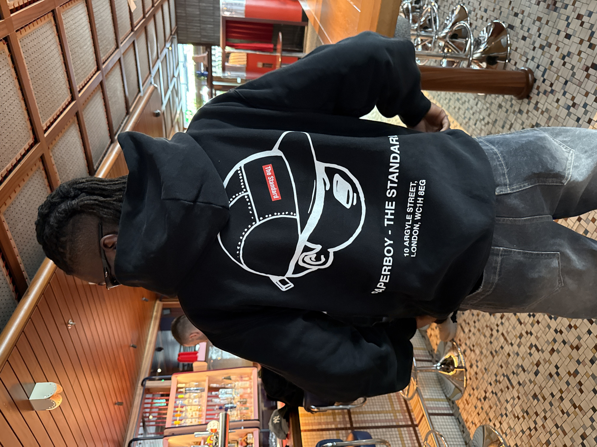 A person is depicted from behind, standing or sitting in an indoor commercial space, likely a hotel, bar, or restaurant. The individual wears a black hooded sweatshirt and dark grey jeans. The hoodie features a prominent white graphic design on the back, illustrating a stylized head wearing a cap and sunglasses. A small red rectangular label on the cap within the graphic reads "The Standard" in white text. Below the graphic, large white text states: "PAPERBOY - THE STANDARD," followed by the address "10 ARGYLE STREET, LONDON, WC1H 8EG."
The person has dark hair styled in dreadlocks or braids, visible at the back of their head and neck. The background features wooden paneled walls with light-colored woven or cane inserts on the left side of the frame. The floor on the right consists of small, mosaic-like tiles in varying shades of brown, beige, and grey. Chrome bar stools with dark seats are partially visible in the foreground. In the mid-ground, a decorative metallic object resembling curved horns or tubes is mounted above a wooden base. The overall lighting suggests a well-lit interior, indicative of daytime or a brightly illuminated evening setting. The visible address confirms the location as The Standard hotel in London, United Kingdom.