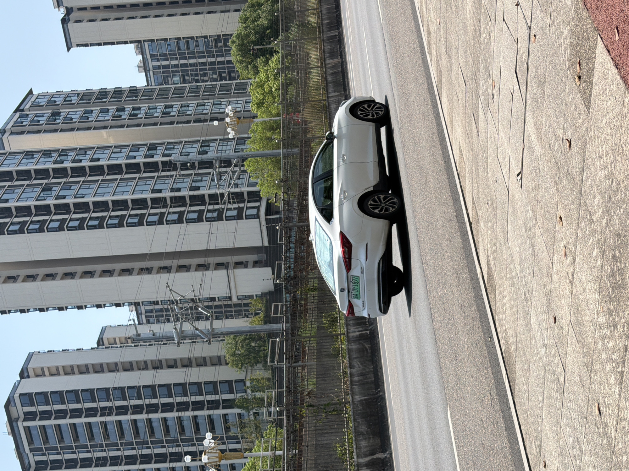 The image captures a street scene in Jiangjin, China. In the foreground, a white sedan with a license plate reading "A-DY1897" is traveling on a paved road. The car is viewed from the rear-right quarter, with its left rear tire on the asphalt. To the left of the road, a sidewalk made of large, rectangular stone tiles is visible. Beyond the road, a concrete barrier is topped with a fence, behind which lush green trees and foliage create a natural buffer. In the background, towering apartment buildings with numerous windows dominate the skyline, indicative of a dense urban environment. The sky above is a clear, pale blue. The overall impression is a snapshot of everyday urban life in a Chinese city.