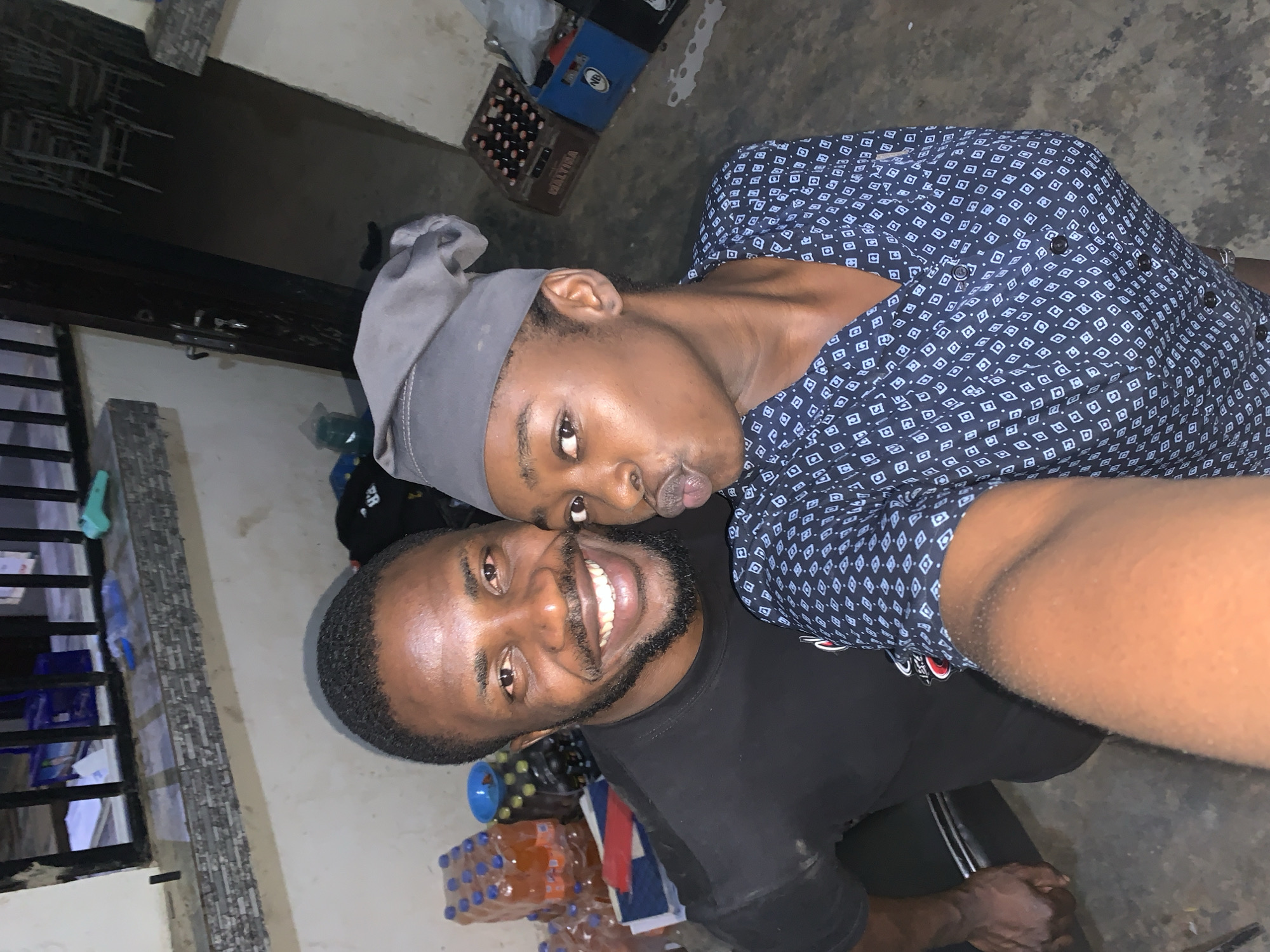 Two young African men are posing closely for a selfie in what appears to be a residential or commercial space in New Stadium, Nigeria. The man in the foreground wears a black t-shirt and sports a beard and mustache. He is smiling broadly, showcasing his teeth. The second man, slightly behind, is wearing a patterned blue and white shirt and a gray head covering. He is pursing his lips as if to give a kiss. In the background, visible objects include cases of soft drinks and beer, which are common items in commercial areas. The lighting is dim, suggesting an indoor setting, possibly at night. The environment is somewhat cluttered, with items stored around the edges of the space. The walls appear to be painted a light color with some tiling or decorative stone-like elements.