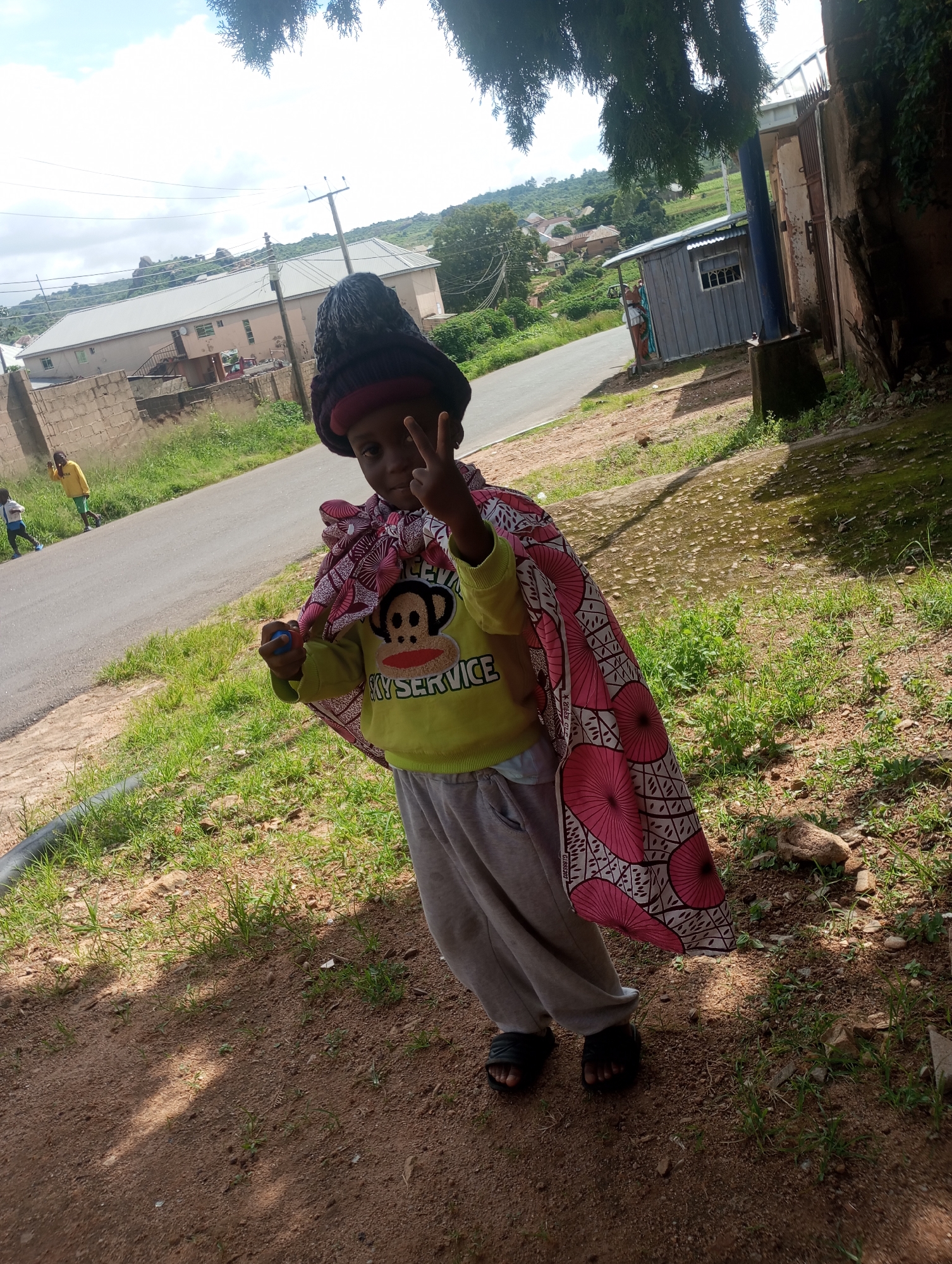 A child wearing a black knit cap, a yellow long-sleeved shirt with a monkey graphic and "SKY SERVICE" text, and gray sweatpants stands on a dirt path in front of a paved road. The child is wrapped in a patterned pink and white cape and holds a small blue object. The child is making a peace sign with their right hand. In the background, there is a beige building with a metal roof, a wooden shed, a utility pole, and lush green hills with scattered buildings. The ground is covered with dry dirt, sparse green grass, and rocks. The image appears to have been taken in Jos, Nigeria, given the distinctive architecture and landscape often found in the region.