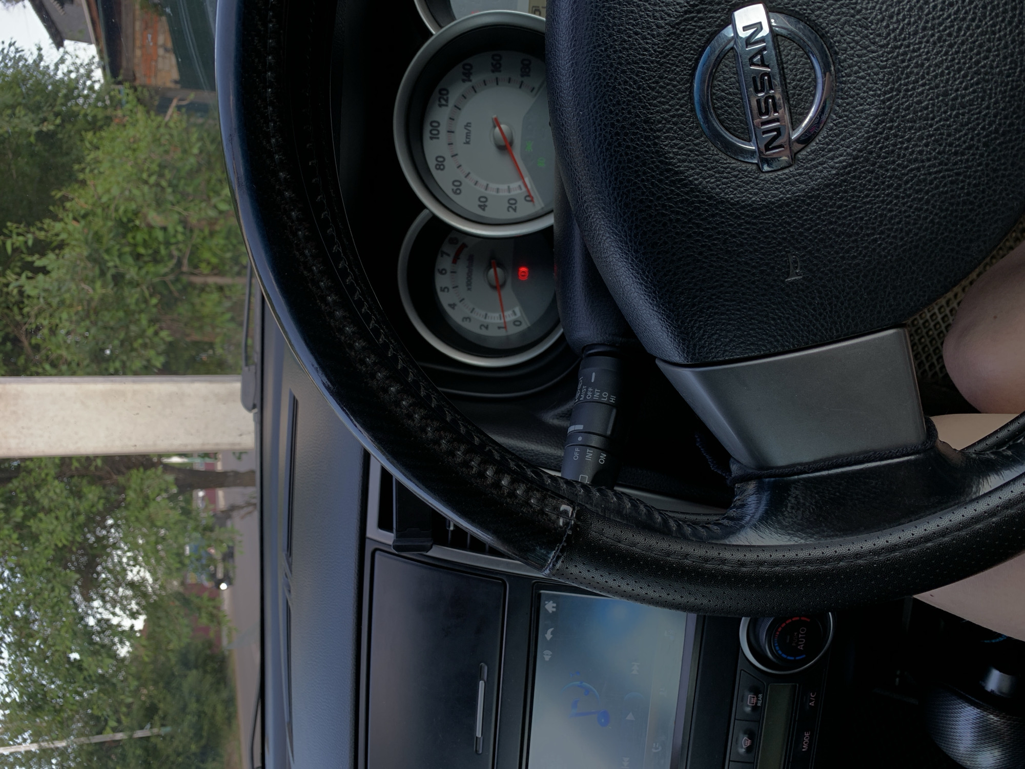The image shows the interior of a Nissan car, specifically the dashboard and steering wheel. Outside the car, through the windshield, a rural or suburban road is visible with trees and a building in the background. The car is likely stationary, as indicated by the unmoving gauges. The steering wheel is black leather with the Nissan logo in the center. To the left of the steering wheel is the car's infotainment system, displaying a music interface. Below that, there are climate control buttons and a gear shifter. The overall impression is that of a typical car interior on a slightly overcast day, possibly in a temperate climate. The specific mention of Belogorsk, Russia is not directly verifiable from the visual information alone, but if this context is provided, it suggests the car is located there.