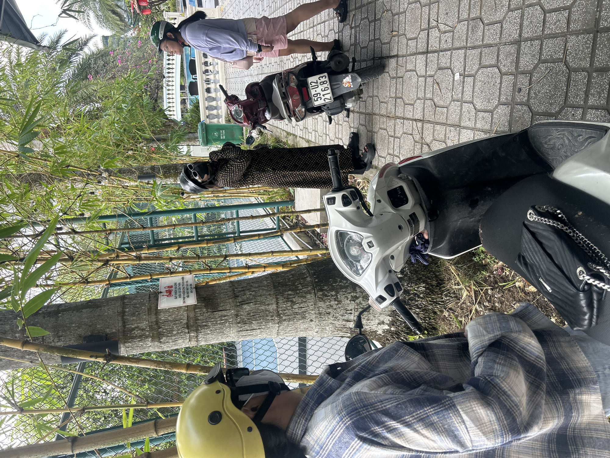 In Thanh Hưng, Vietnam, two individuals are standing near two parked scooters. The person on the left, wearing a yellow helmet and a blue and white plaid shirt, is partially visible. They are facing towards the right side of the image. The person on the right is wearing a green helmet and a light purple hoodie with pink shorts, looking towards a swimming pool area in the background. The second person, standing closer to the center of the image, is wearing a dark patterned long dress, a dark helmet, and sunglasses, with their hands clasped in front of them.
The environment includes a paved walkway with gray hexagonal and square tiles. Behind the people and scooters, there is a tall tree with a sign attached to it. The sign has Vietnamese text and an illustration of a scooter. Bamboo stalks are visible behind the tree and along the fence line. A green trash bin is also present. In the background, a white railing surrounds a pool, and palm trees with some pink flowers are visible. One scooter is dark red, and the other is white. The license plate on the red scooter reads "59-U2 216.83".