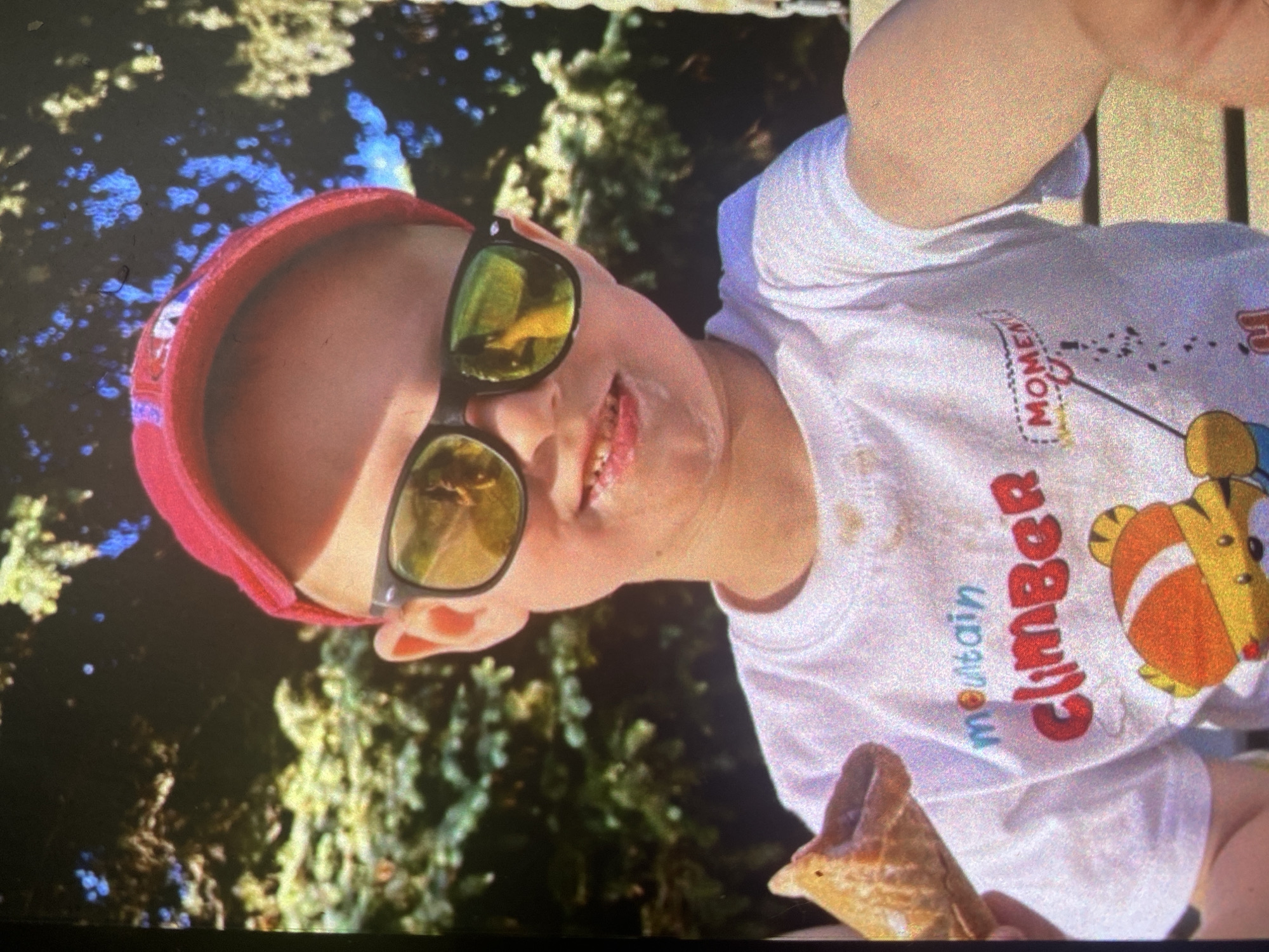 An outdoor, close-up portrait, originally oriented vertically, captures a cheerful young child. The child wears a pink or red headband and black-framed sunglasses with reflective yellow-green lenses. They are dressed in a white short-sleeved t-shirt that prominently features the text "mountain climber" in red and blue, with the word "MOMEN" also visible and a graphic of a cartoon animal, possibly a tiger cub, rendered in orange and yellow. The child's mouth is slightly open, revealing teeth and some light-colored residue around the mouth, suggesting a recent treat. In one hand, they hold a partially eaten waffle ice cream cone. The background is a blurred expanse of green foliage, indicating a sunny outdoor setting such as a park or garden. The bright, clear lighting suggests it is daytime. The child appears happy and is actively enjoying their ice cream. The provided context suggests the location is Saint Petersburg, Russia, though specific visual cues for this location are not present in the image.