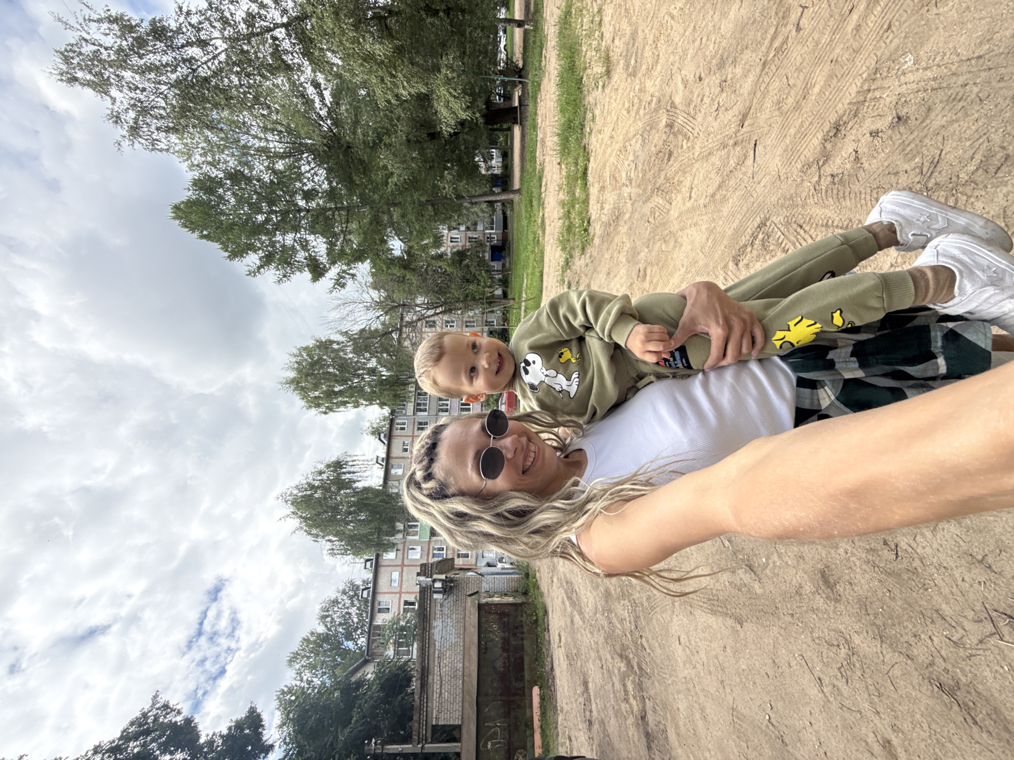 An outdoor, eye-level, medium shot captures a woman holding a young child. The woman, with blonde hair styled in braids and wearing sunglasses, smiles at the camera. She is dressed in a white tank top and a plaid skirt. The child, wearing a green tracksuit with a Snoopy graphic, looks to the side. They are positioned on a dirt ground, with apartment buildings and trees in the background. The scene appears to be in Rusanovo, Russia, a rural village within the Tver Oblast, approximately 30 kilometers from Tver city. The weather is cloudy.