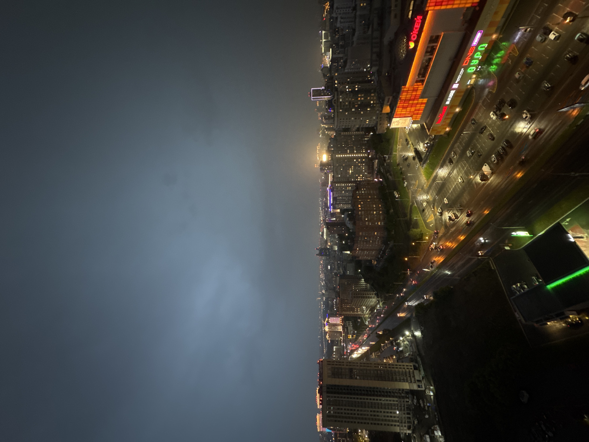 This is a nighttime aerial view of a city scene, likely Novosibirsk, Russia, characterized by a dark, overcast sky with subtle hints of distant lightning. The foreground is dominated by a wide, wet road reflecting the colorful lights of the city and passing vehicles. On the right side of the road, a brightly lit commercial building with large illuminated signs, including "DNS" and "О'КЕЙ," stands out. Adjacent to this building is a large, illuminated parking lot filled with numerous cars. Across the road, several tall residential or office buildings with illuminated windows are visible, some featuring colorful architectural lighting. In the mid-ground and background, a sprawling cityscape extends into the distance, with many buildings lit up, and the overall atmosphere is moody and illuminated by artificial light against the dark sky.