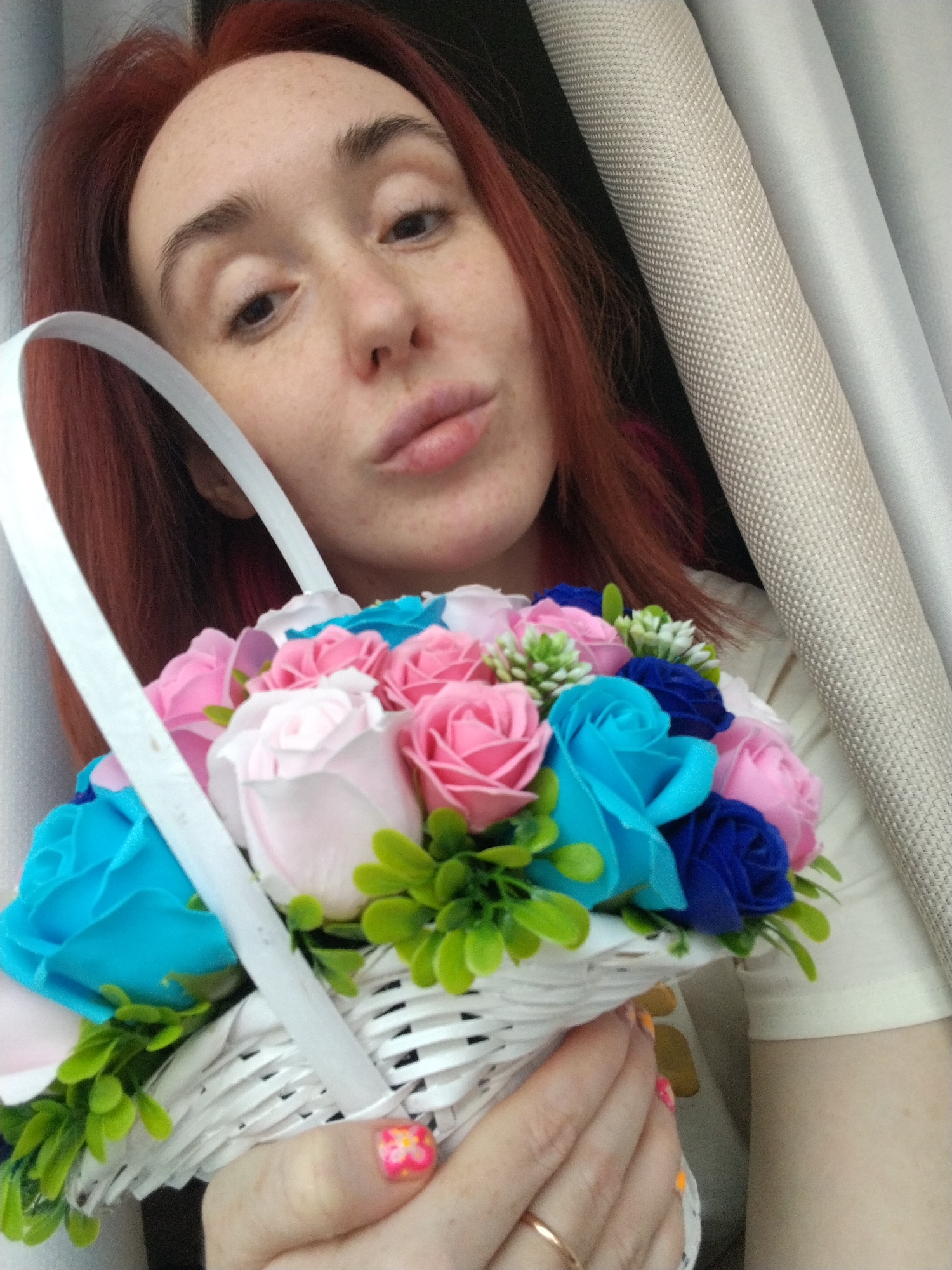 An adult female, characterized by red hair, freckled fair skin, and dark eyes, is depicted in a close-up, high-angle shot. Her lips are slightly pursed as she looks directly into the camera. She holds a white woven basket filled with artificial flowers, including light pink, dark pink, light blue, and royal blue roses, accented by green foliage. Her left hand, visible in the lower right, wears a gold-toned ring on the ring finger, and her fingernails display a floral manicure. She is wearing a light-colored short-sleeved garment. The background consists of draped fabrics: a textured light beige material on the right and a smoother light-toned fabric next to it, with a dark area visible on the far left. The image provides no visual information to ascertain a specific location within Izhevsk, Russia.