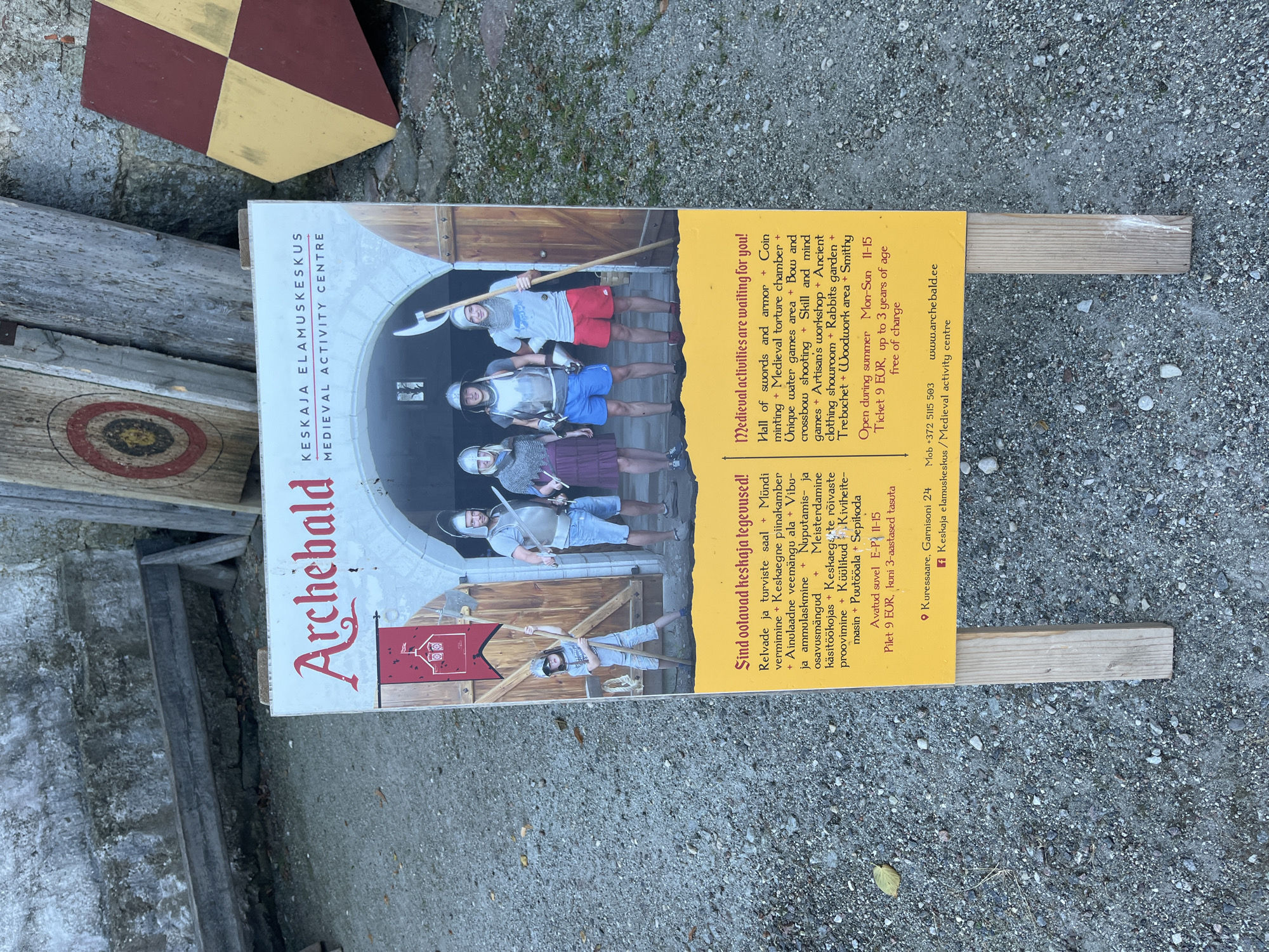 The image shows a sign for the "Archebald Medieval Activity Centre" located in Kuressaare, Estonia. The sign is a bright yellow with red text and features a photograph of four individuals in medieval-style armor and clothing standing in an arched doorway.
The text on the sign lists various activities offered at the center, including: Hall of swords and armor, Coin minting, Medieval torture chamber, Unique water games area, Bow and crossbow shooting, Skill and mind games, Artisan's workshop, Ancient clothing showroom, Rabbits garden, Trebuchet, Woodwork area, and Smithy.
The sign also indicates that the center is open during the summer from Monday to Sunday, 11:00 AM to 3:00 PM. The ticket price is 9 EUR, and children up to 3 years old are admitted free of charge. The location is specified as Kuressaare, Garnisoni 24. Contact information includes a mobile number (+372 5115 503) and a website (www.archebald.ee). The bottom of the sign also displays the Estonian text "Keskaja elamuskeskus / Medieval activity centre".
The sign is propped up on two wooden stands in an outdoor setting. The ground surface appears to be gravel and dirt. In the background, there are wooden targets and a stone wall, suggesting the location is an outdoor attraction or historical site.