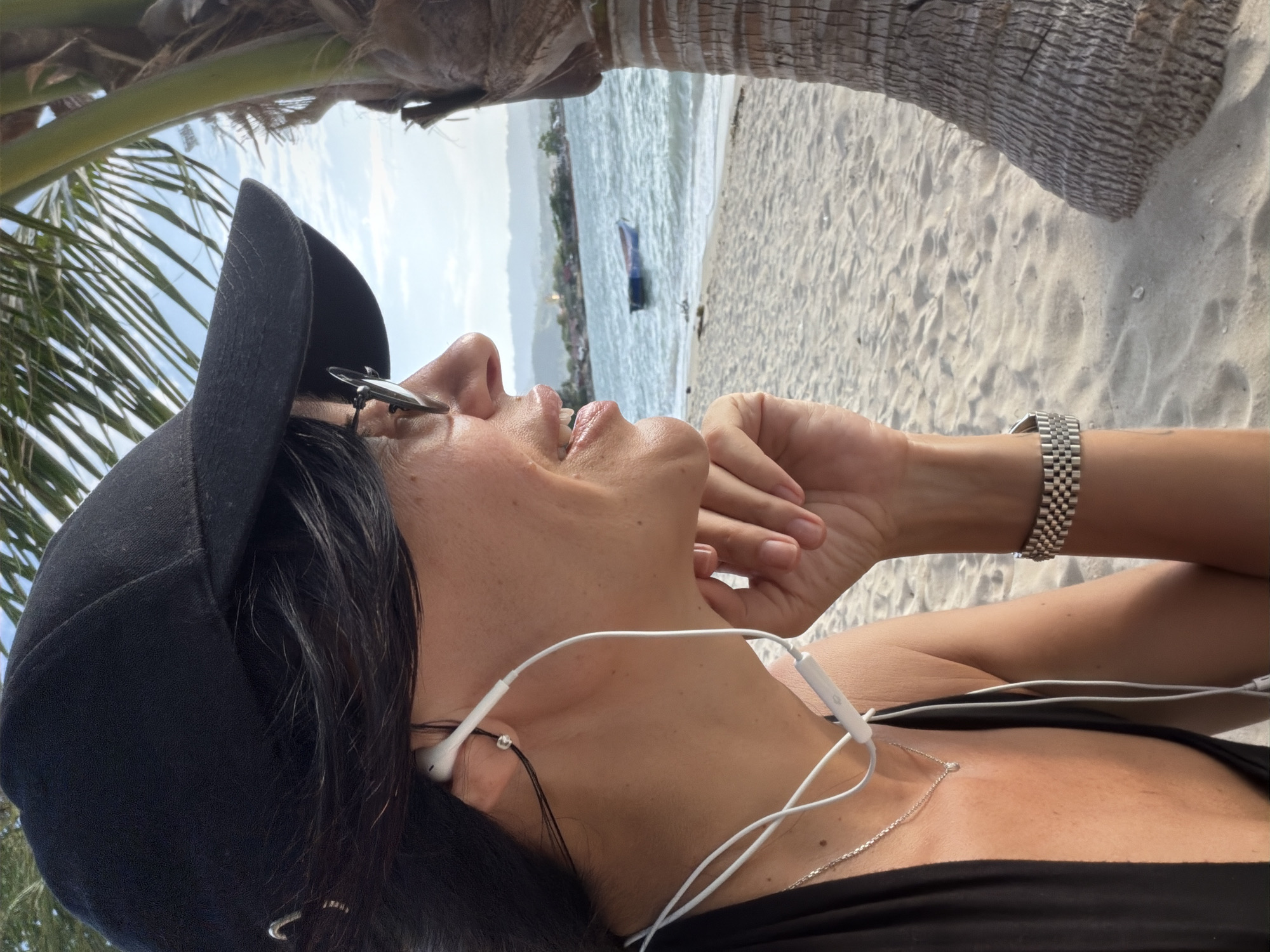 A medium shot captures a woman in profile, wearing a black baseball cap, sunglasses, and earbuds, looking out at a body of water. She has dark hair and is wearing a black top. A silver necklace with a small pendant is visible on her chest. Her right hand is raised to her chin, and she wears a silver watch on her left wrist. The background consists of a sandy beach, palm trees, and the sea with a small boat and distant landmass. The lighting suggests it is daytime, possibly late afternoon, with a soft, warm quality. The scene appears to be a tropical beach setting.