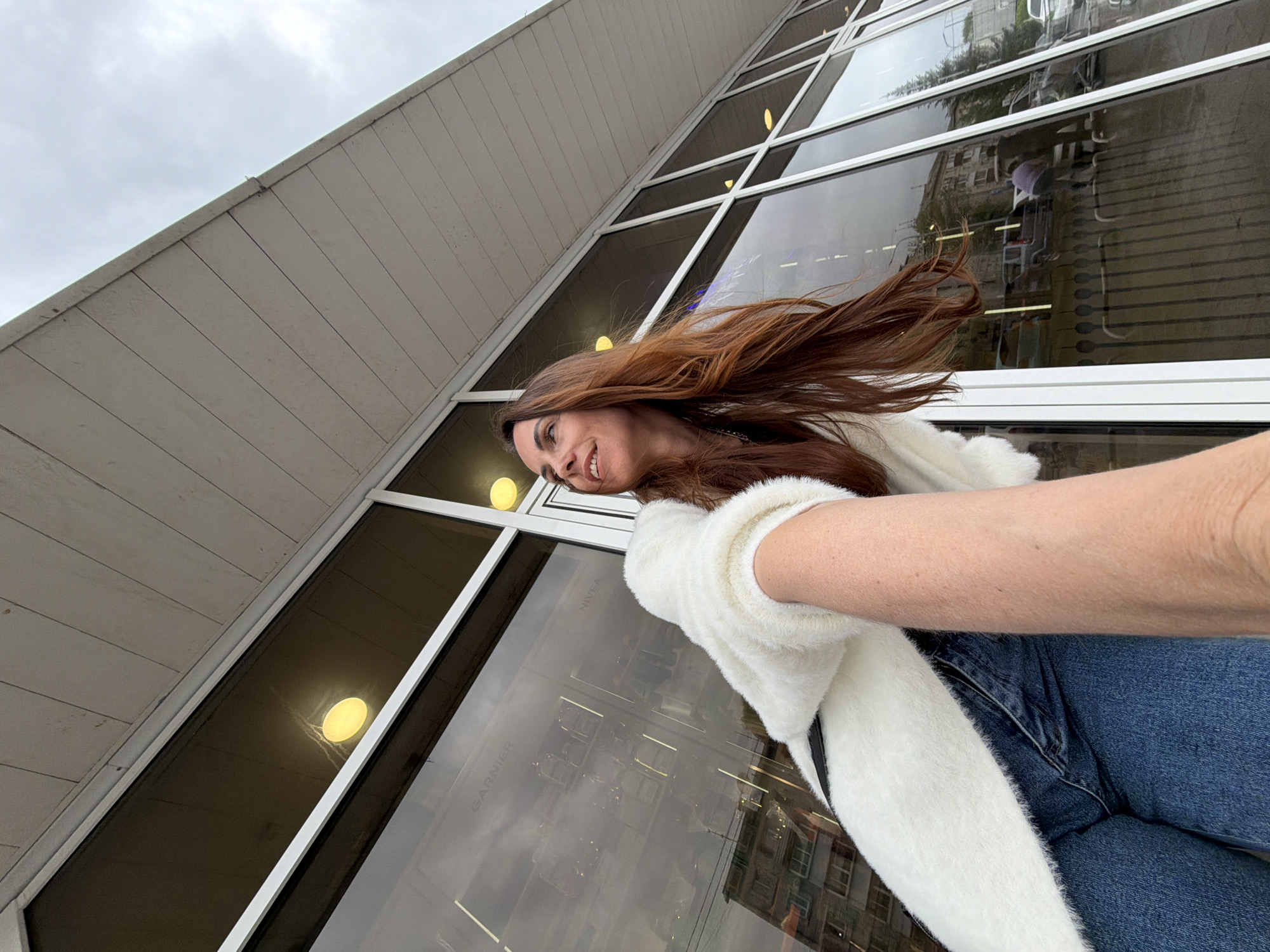 A low-angle shot depicts a woman taking a selfie in front of a building. The woman has reddish-brown hair that is blowing in the wind, and she is wearing a white, fluffy cardigan and blue jeans. The building behind her has a row of large, rectangular windows with white frames. The windows reflect the surrounding environment, including other buildings and the sky.
The reflections in the windows suggest that the location might be a commercial area, potentially a shopping street. The presence of visible reflections within the windows makes it difficult to identify the specific building. It is not possible to ascertain the city of Birobidzhan, Russia from the image.