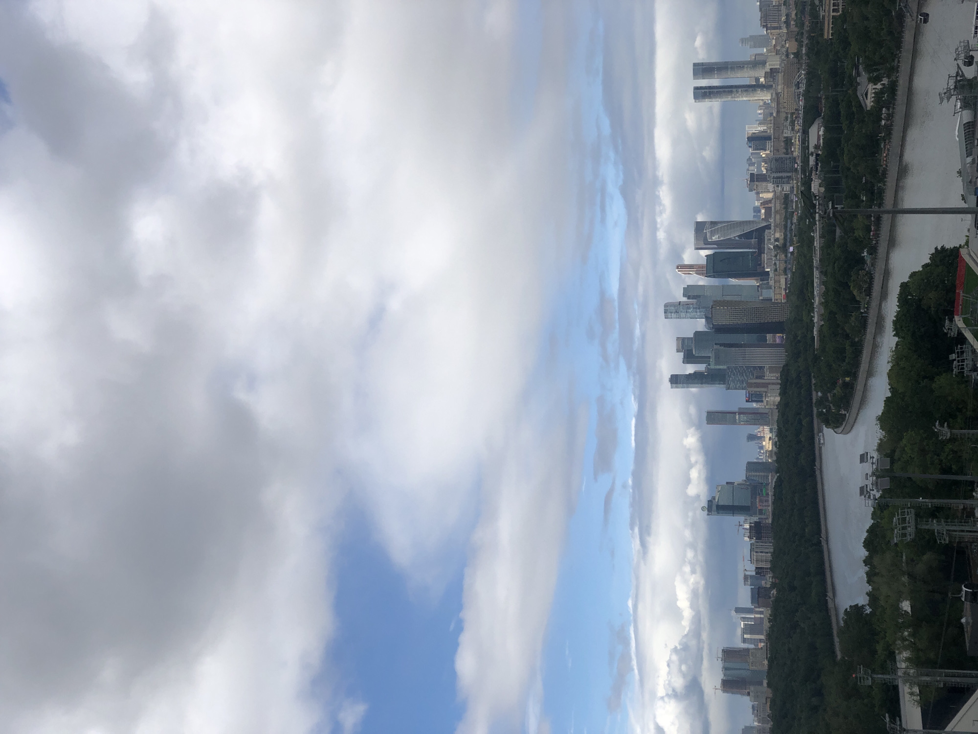 The image shows a view of the Moscow International Business Center (Moscow City) in Moscow, Russia. The photo is taken from an elevated position, looking towards the skyscraper complex. In the foreground is the river, the embankment, and greenery. On the right, there are some utility poles. The sky is partially covered with clouds.