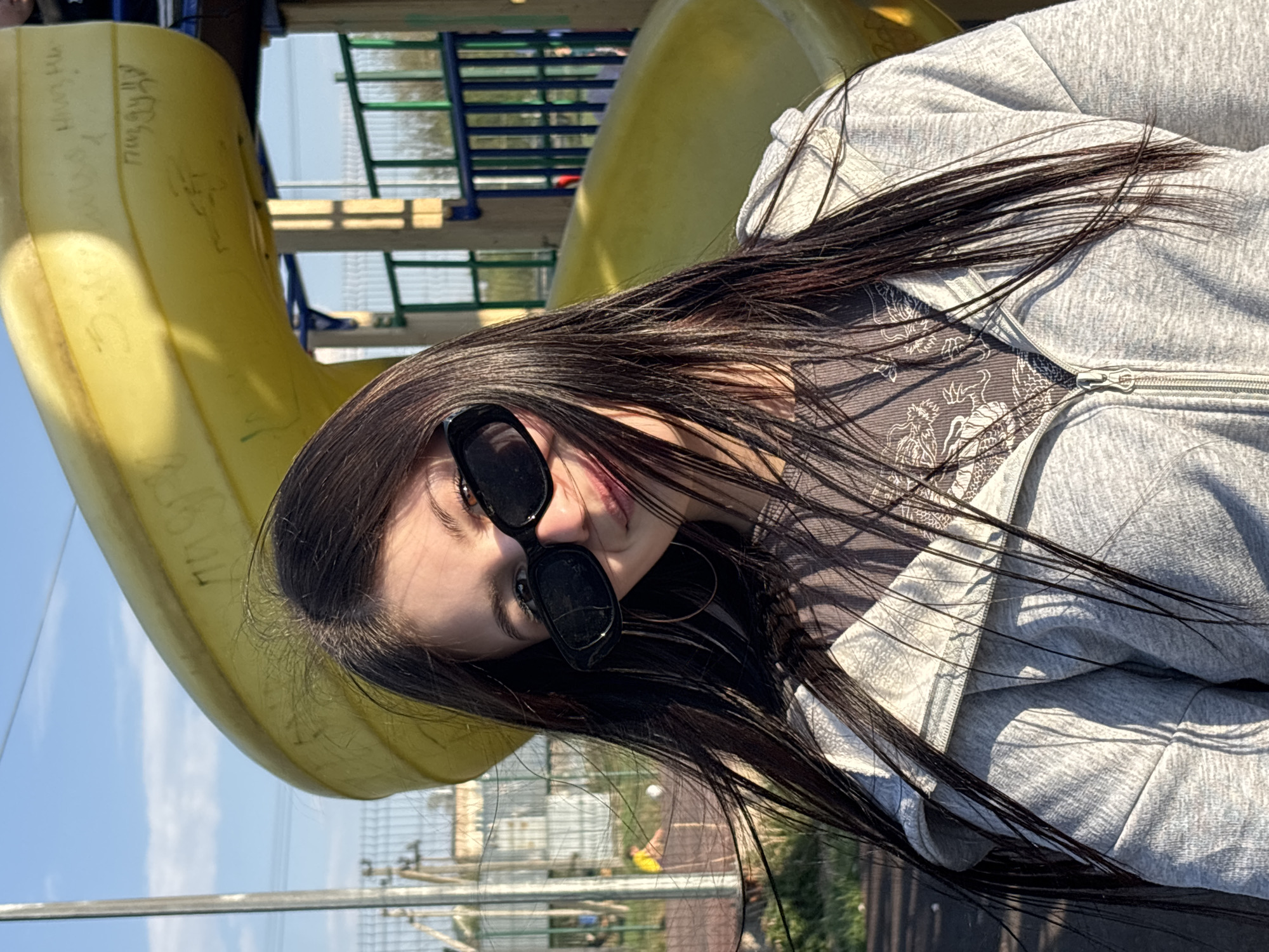 A young woman with long, dark hair and sunglasses is posing for a photo outdoors, likely at a playground in Фоки, Russia. She is wearing a grey zip-up hoodie. She is sitting or leaning against a bright yellow slide. The background includes playground equipment and a view of the sky.