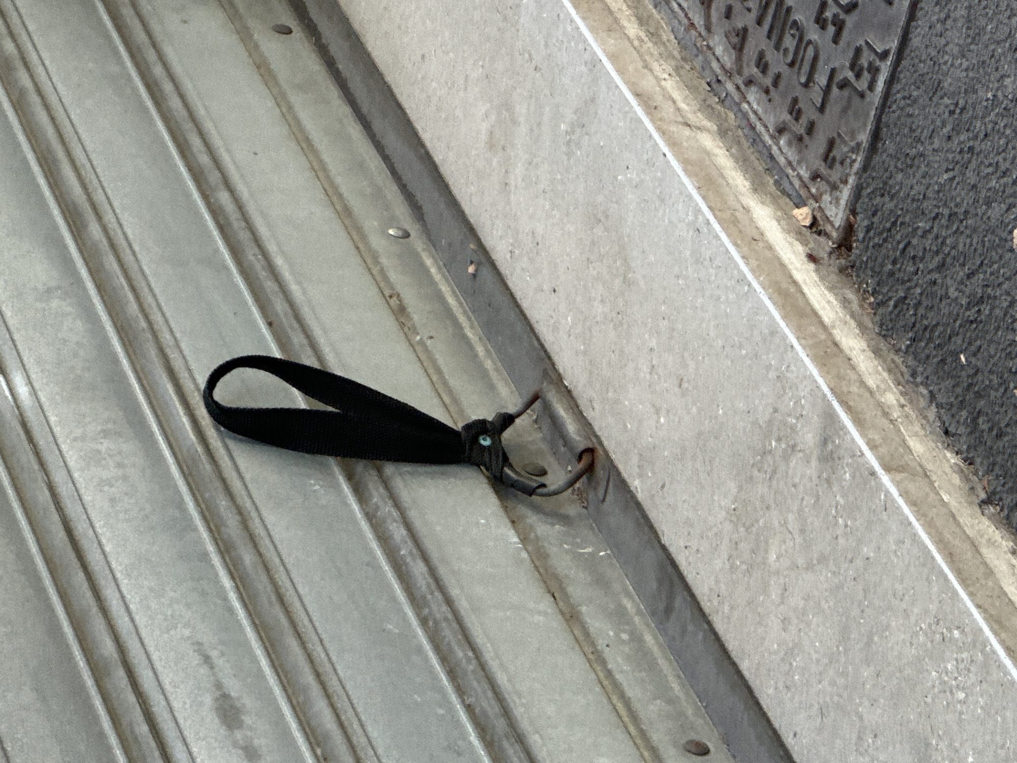 A black fabric loop is attached to a metallic, corrugated shutter. The loop is secured with a dark green string or cord, which is knotted around a dark metal hook or clasp that protrudes from the shutter. The shutter appears to be part of a shopfront or garage door. In the foreground, a concrete step or ledge runs horizontally across the image. To the right of the step, a textured dark surface, possibly asphalt, is visible, with a metal grate or manhole cover embedded in it, displaying partial lettering that reads "FOGNA". The overall scene suggests an outdoor urban environment in Trieste, Italy.