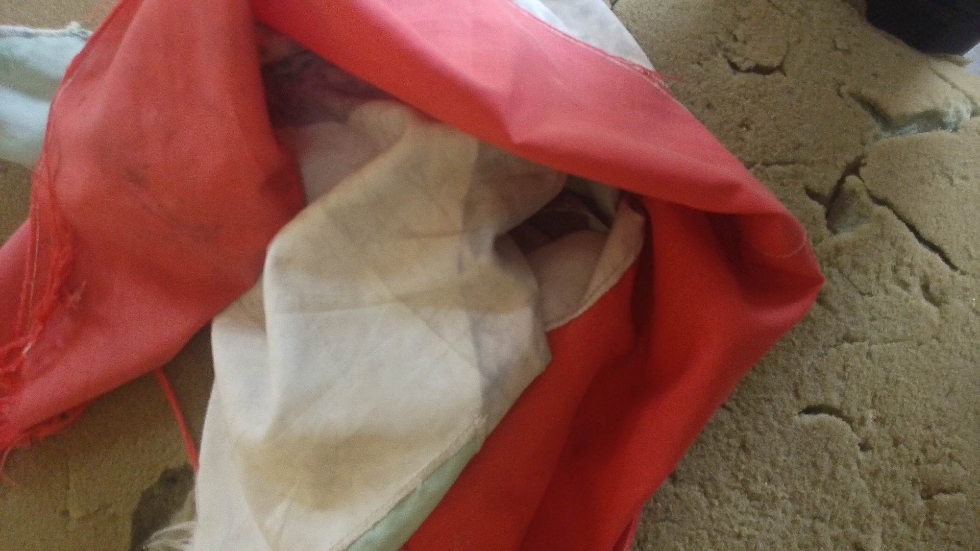 The image shows a close-up view of a crumpled red and white fabric, possibly a flag or banner, lying on a textured, light brown surface that appears to be cracked and somewhat degraded. The fabric is bunched up, with the red material dominating the foreground and upper portion of the image, while a lighter, off-white fabric is visible beneath and to the left. The red fabric has a frayed edge visible on the left side. The brown surface has an irregular texture with visible cracks and depressions, resembling weathered concrete or a similar composite material. There are no people or discernible actions taking place. The lighting suggests an indoor or shaded outdoor environment. The image does not provide any specific details that would allow for the localization of this scene within the city of Balewa, Nigeria.