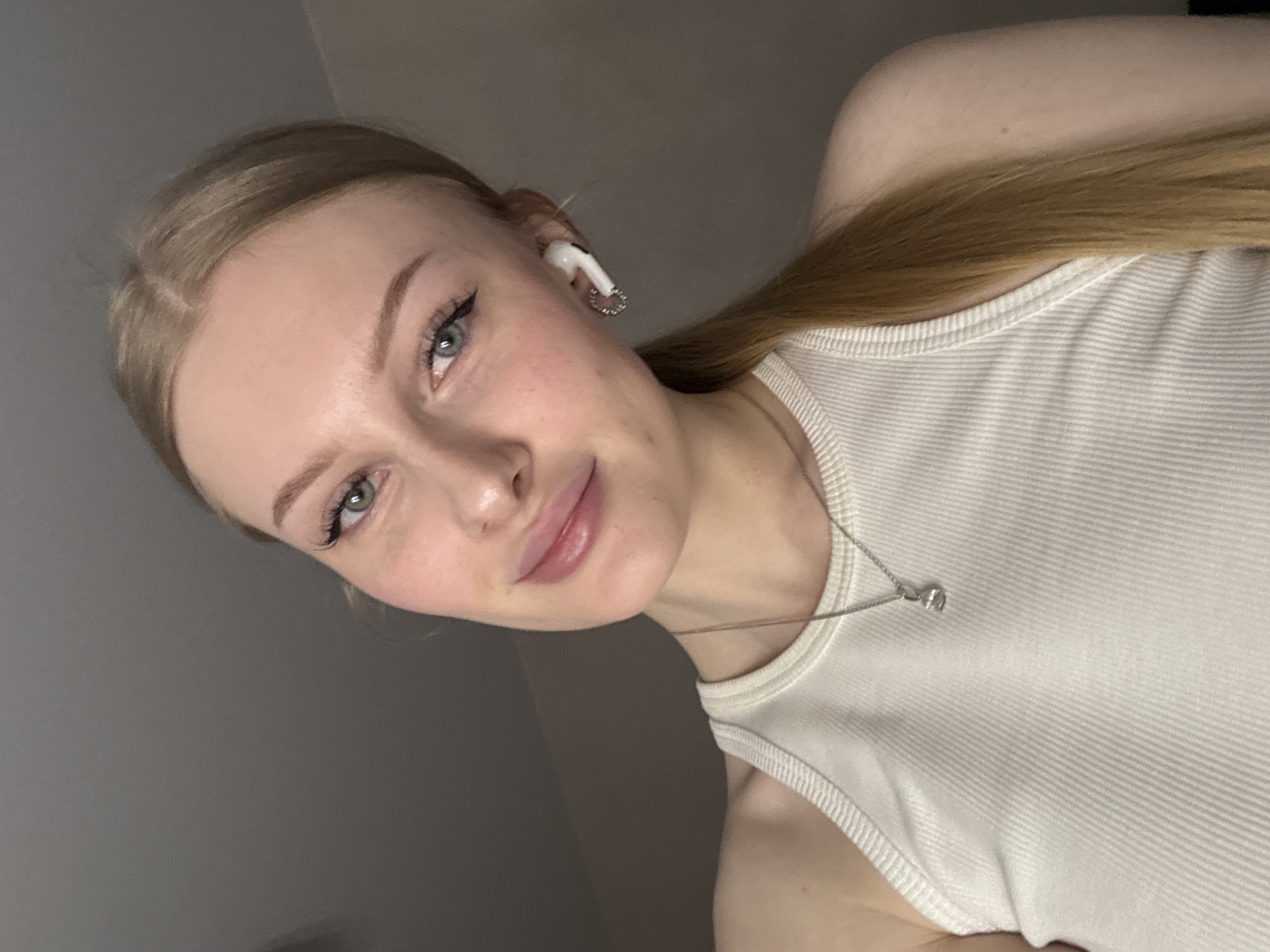 A fair-skinned young woman with light brown hair pulled back in a ponytail is facing the camera. She has light-colored eyes, subtly defined eyebrows, and pink lips. She is wearing a white ribbed tank top and a silver necklace with a small pendant. A white earbud is visible in her left ear. The background appears to be a grey wall. There is no evidence of location, so providing a location in Perm, Russia, is not possible.