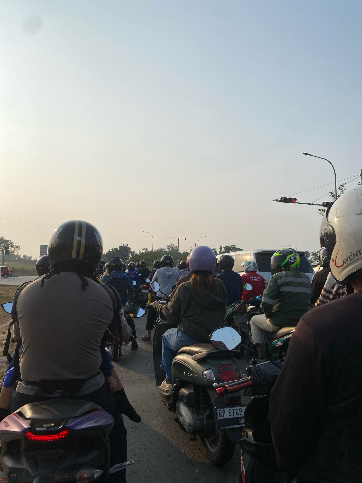 The image depicts a traffic jam of motorcycles on a road in Batam, Indonesia, likely during daylight hours, given the bright, hazy sky. Numerous individuals, predominantly wearing helmets for safety, are positioned on their motorcycles, forming a dense queue. Some riders are in motion, indicated by the blurred backgrounds and slight movements of the vehicles, while others appear to be stationary, waiting for traffic to clear. The environment features paved roads, flanked by what appears to be undeveloped land with sparse vegetation. In the distance, there are hints of urban infrastructure, including traffic lights and street lamps, suggesting the scene is taking place within or near a developed area of the city. The presence of various motorcycle models and rider attire suggests a mix of personal transportation.