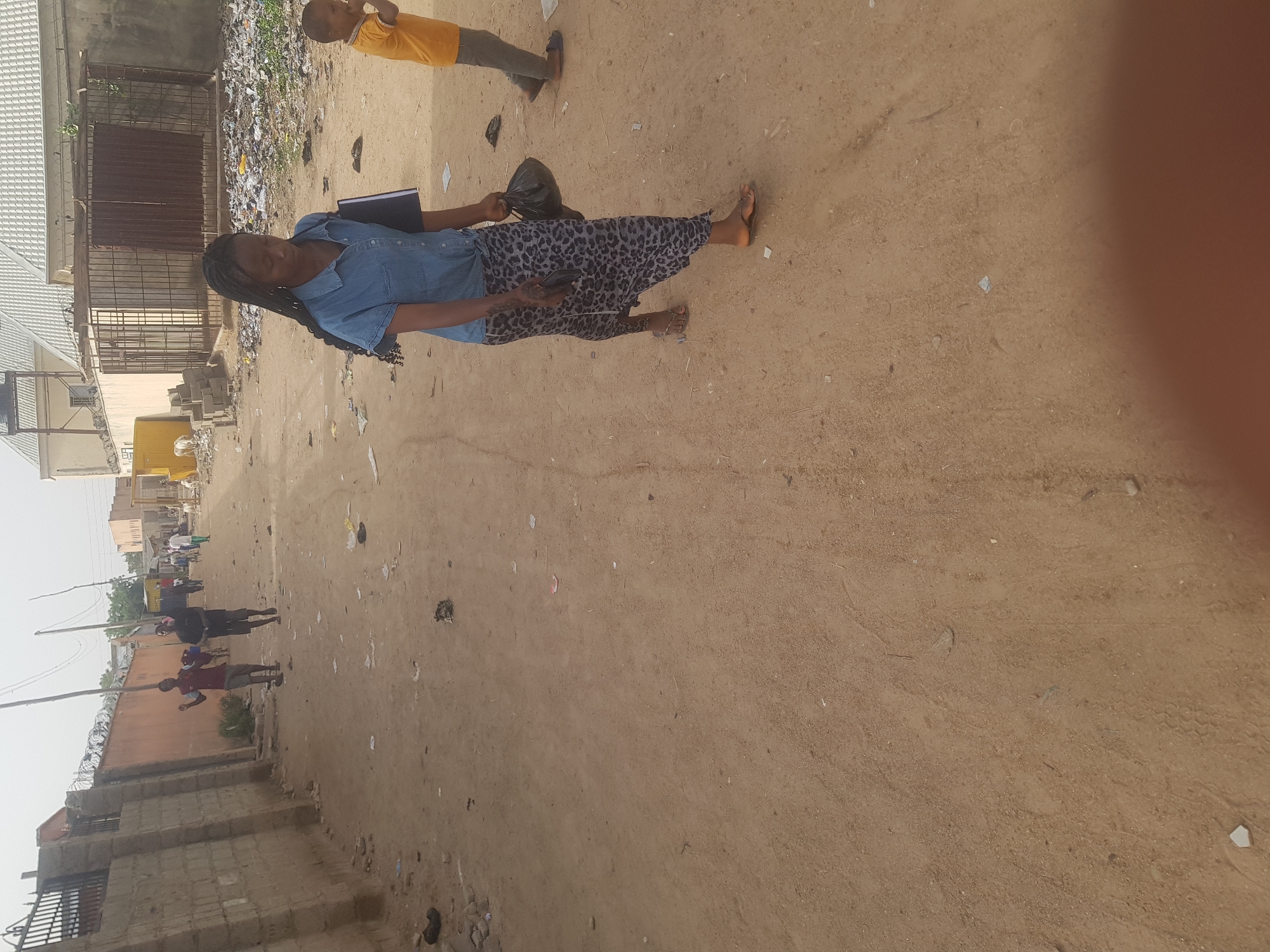 The image captures a street scene in Dabawa, Nigeria. A woman with long braids is centrally located, wearing a light blue top and patterned pants. She carries a dark-colored bag and a book or folder. A child in a yellow shirt stands slightly behind her. The street is unpaved and sandy, scattered with debris. Buildings line both sides of the street, with a mix of concrete block structures and metal fencing. Other figures are visible further down the street, appearing to be walking. The sky is overcast, casting a relatively even light across the scene.