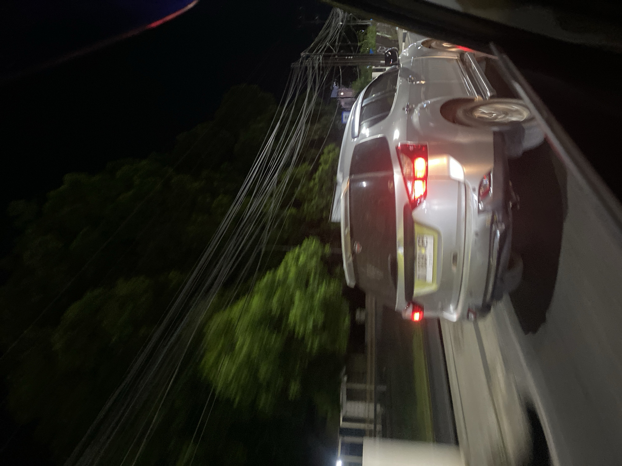 The image shows the rear of a silver SUV at night in Sungai Paring, Indonesia. The vehicle is parked on a paved road, with dense green trees on the left side of the frame. A tangle of electrical wires hangs precariously over the scene, obscuring some of the background and suggesting a somewhat cluttered or underdeveloped area. The car's taillights are illuminated, indicating it is either stopped or in motion. The lighting suggests it is nighttime, and the darkness of the sky and the illuminated car lights are prominent. There is no visible text in the image. The overall impression is a somewhat ordinary scene of a car at night, with the electrical wires adding a distinctive visual element.