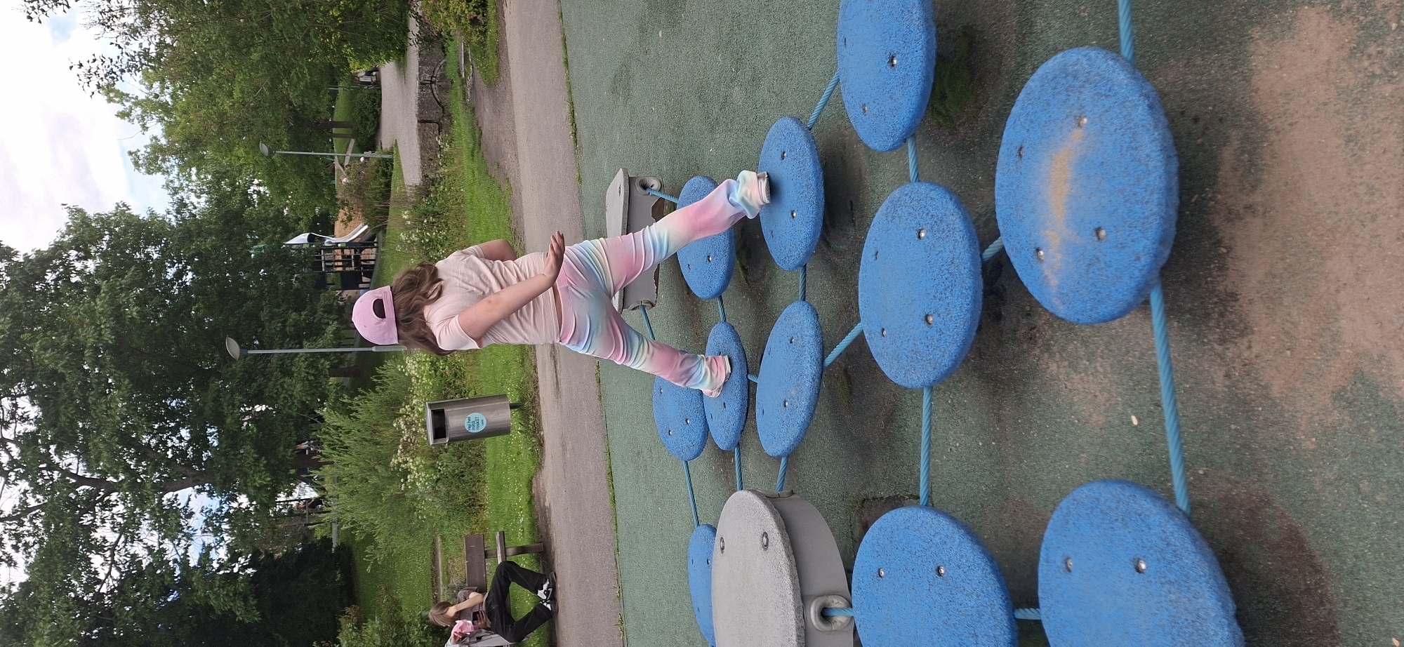 A young girl, wearing a pink baseball cap and pastel tie-dye leggings, is balancing on a playground obstacle course. She is positioned mid-stride between two blue, circular platforms, with her right arm extended backward and her left hand held forward. The platforms are connected by blue ropes and are set on a green, rubberized surface. In the background, a person sits on a park bench, looking at their phone. To the left of the bench is a metal trash receptacle. Further in the background, trees and playground equipment, including a slide, are visible. The scene is set in a park area, possibly in Lahti, Finland, indicated by the presence of typical urban park amenities.