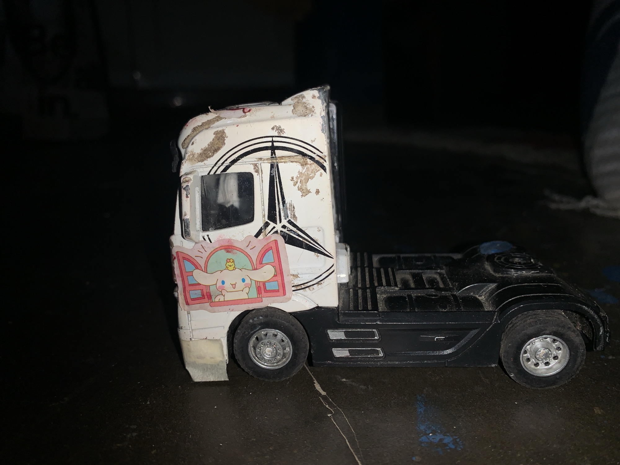 A white and black toy semi-truck cab and chassis is positioned on a dark, slightly reflective surface. The white cab of the truck shows significant wear, with brown, dirt-like residue covering the upper and front portions, and visible paint chipping. A large, black Mercedes-Benz three-pointed star logo is visible on the side of the cab, partially obscured by the dirt and wear. A rectangular sticker featuring the Sanrio character Cinnamoroll, a white dog with long ears, peeking from a pink-framed window with a small yellow chick on its head, is adhered to the lower front side of the cab. The truck's chassis is black, with molded side details. Two silver-colored wheels with black tires are visible: one beneath the cab and one at the rear of the chassis. The background is dark and out of focus, providing no discernible details. The specific location in the city of Karangkemiri, Indonesia, cannot be determined from the visual information presented in the image.