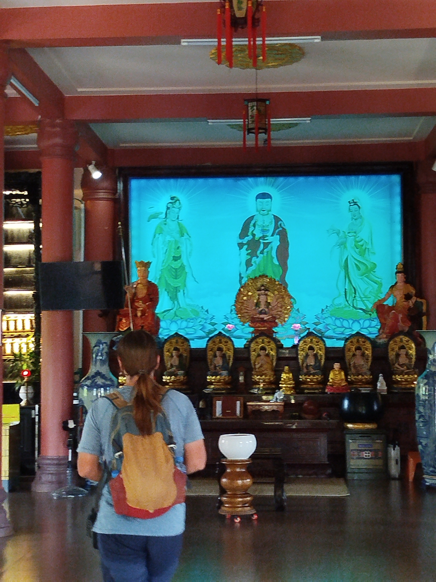 A woman with a backpack stands in a temple in Dien Hong Ward, Vietnam, viewing a large framed image of three figures atop a shelf containing small Buddha statues and various offerings. The temple interior features red pillars and overhead decorations.