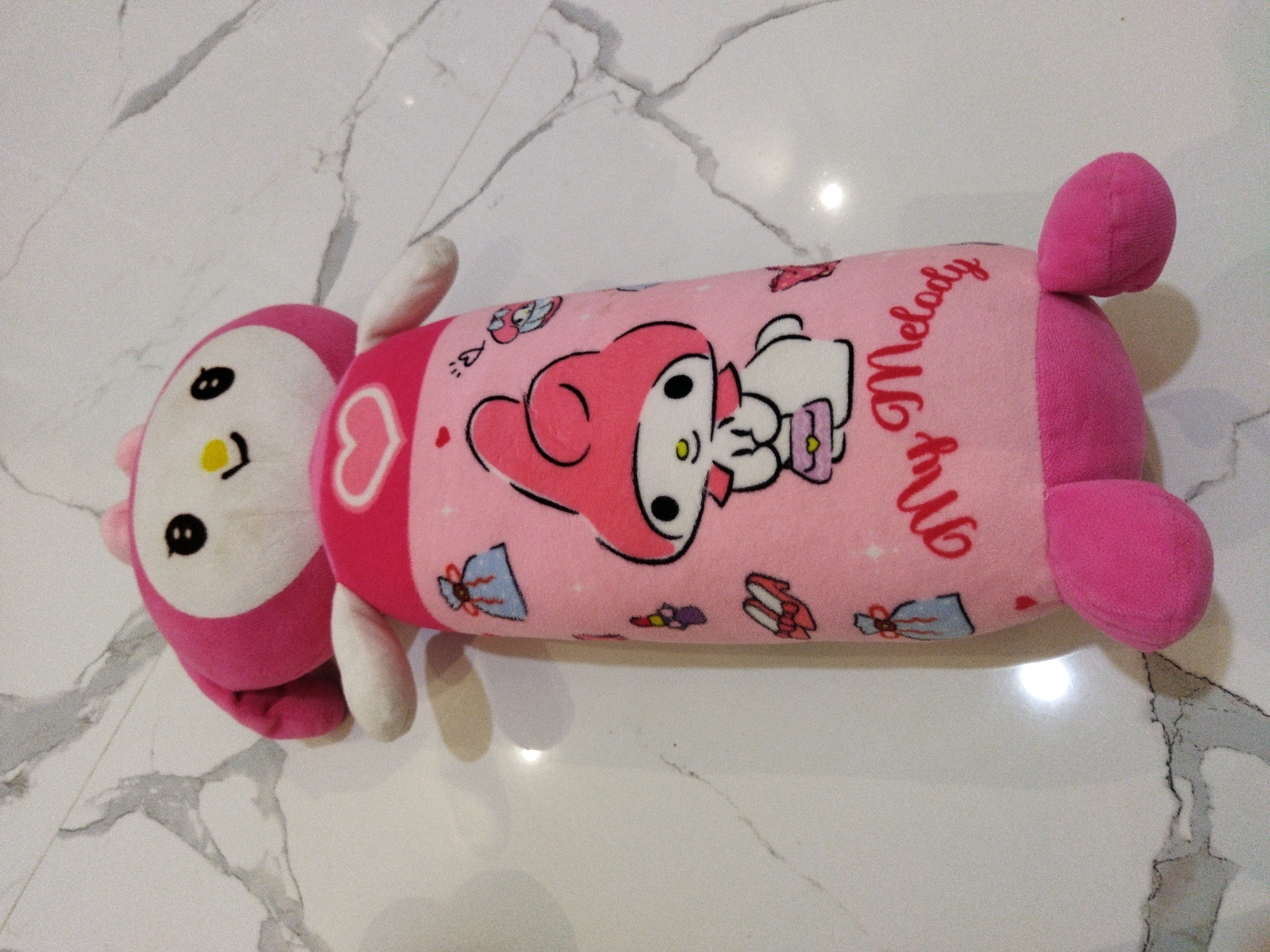 A pink My Melody plush toy is lying on a marble-patterned floor. The toy is cylindrical in shape with a character's face at one end and pink feet at the other. The body of the plush toy is decorated with images of My Melody and bows, and the text "My Melody" is written in cursive red lettering. The plush toy is oriented horizontally in the image, with its head on the left and its feet on the right. The marble floor has a white background with gray veins. The image appears to have been taken indoors. There are no people visible in the image. The location in Banjar Agung, Indonesia is not discernible from the visual information provided in the image.