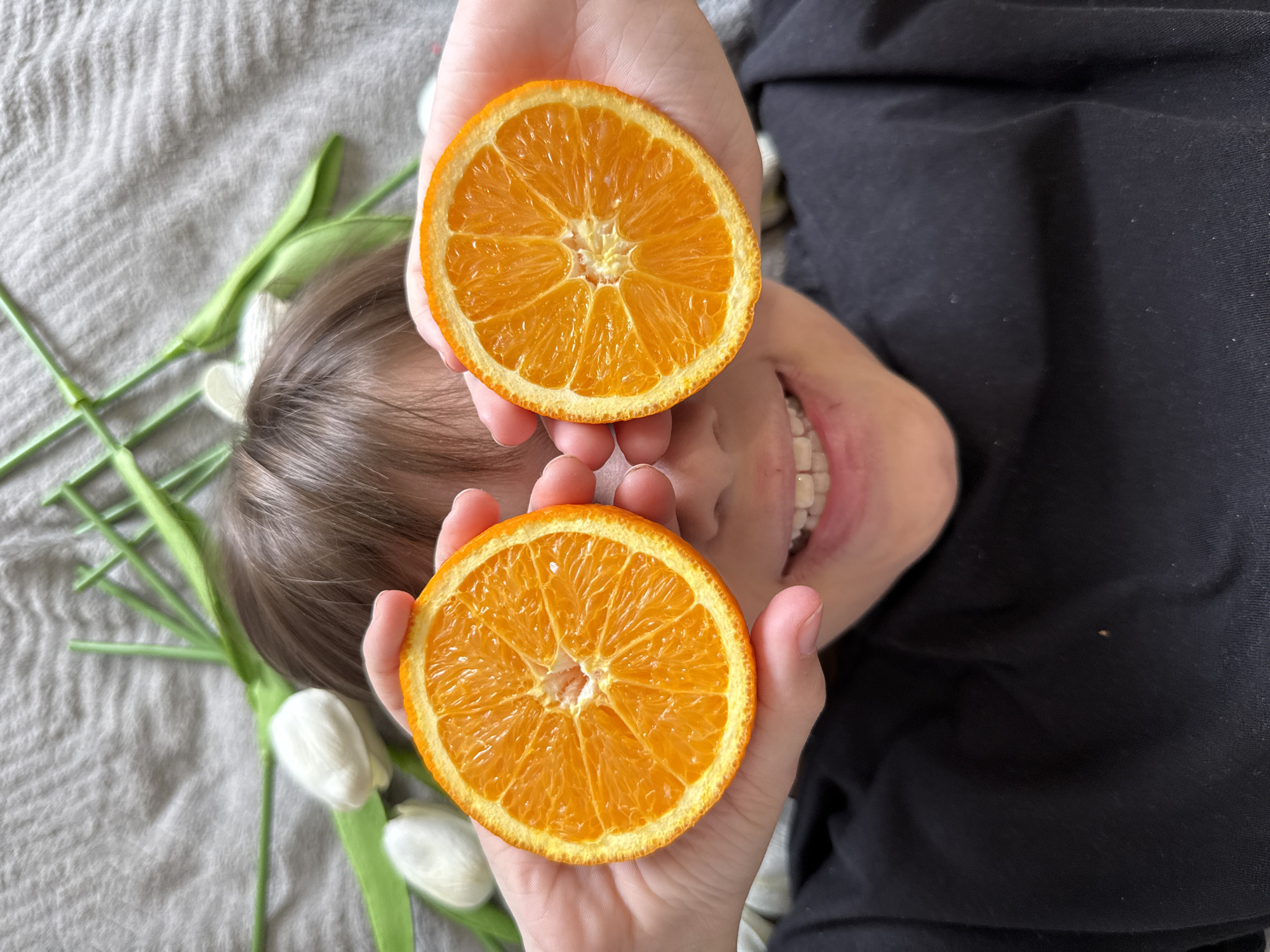 A high-angle shot shows a person lying down on a gray fabric surface, likely a bed or blanket. The person is holding two orange halves directly in front of their eyes. The person is smiling with teeth showing. The person is wearing a black shirt. Several white tulips with green stems are positioned to the left of the person's head.