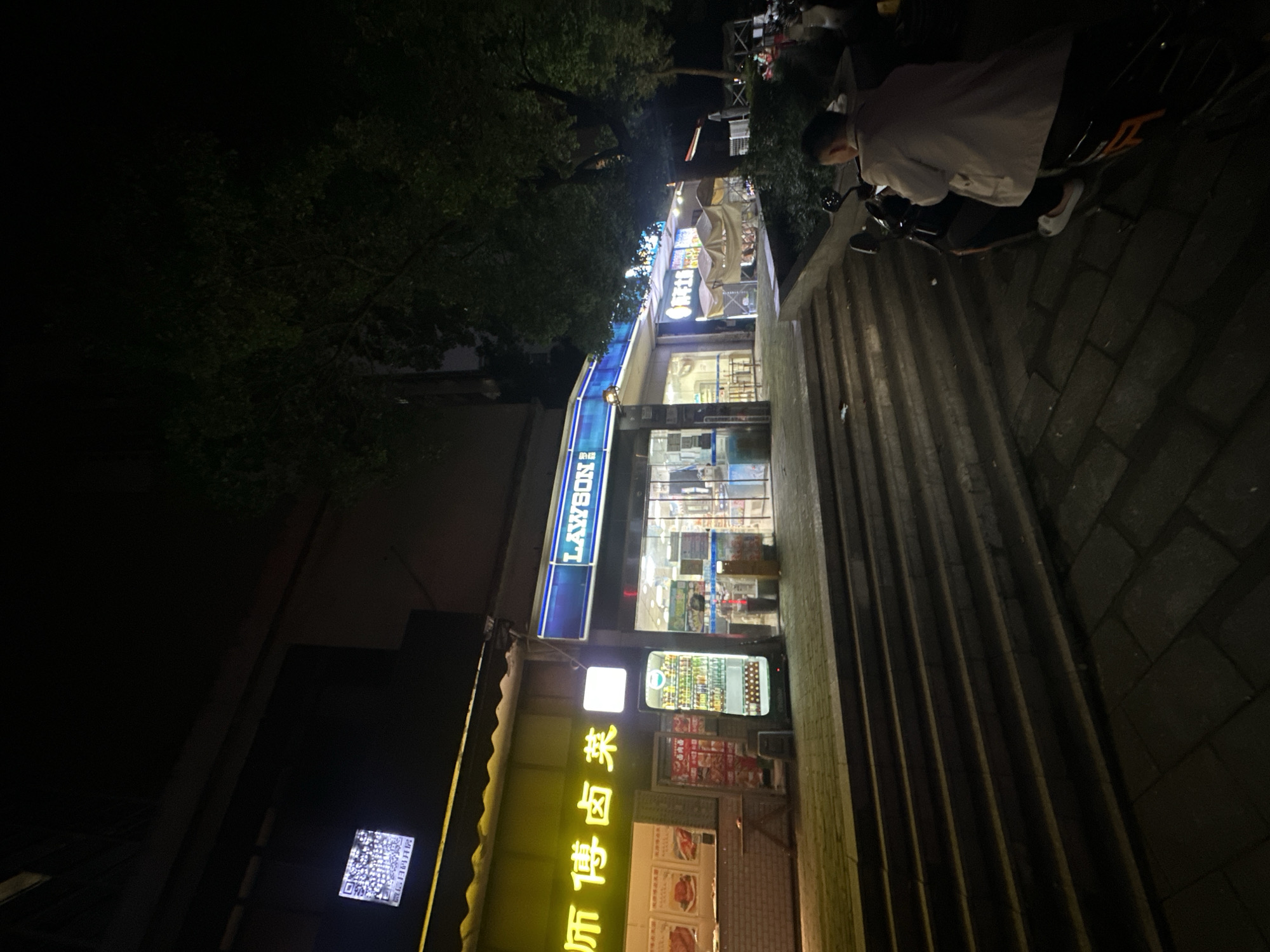 A nighttime photograph depicts a Lawson convenience store in Chongqing High-tech Zone, China. The store is illuminated with bright interior and exterior lighting, showcasing its product displays. Adjacent to the store is a building with a green awning and Chinese characters in yellow neon lights. A person is seated on a scooter or motorcycle, parked on a paved surface near a set of stairs. Trees and foliage are visible in the upper portion of the image. The surrounding environment appears dark, typical of a night scene.