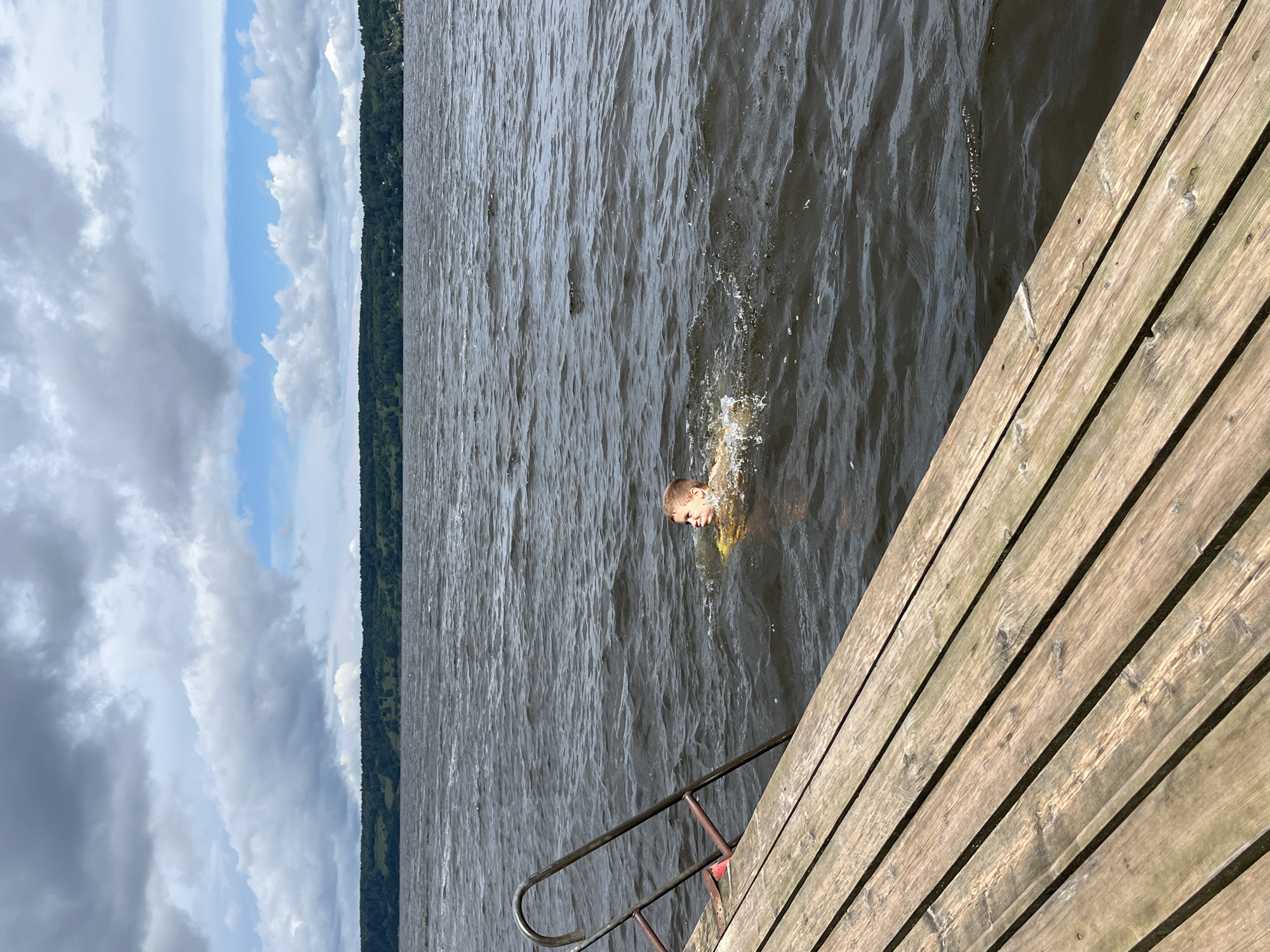 A young boy is swimming in dark, choppy water next to a wooden dock with metal stairs. He is partially submerged and appears to be wearing a yellow flotation device around his waist. The dock is in the foreground, angled from the bottom left corner towards the right. The water extends to the horizon, where a tree-lined shore is visible under a cloudy sky. The location is the city of Lyady, Russia.
