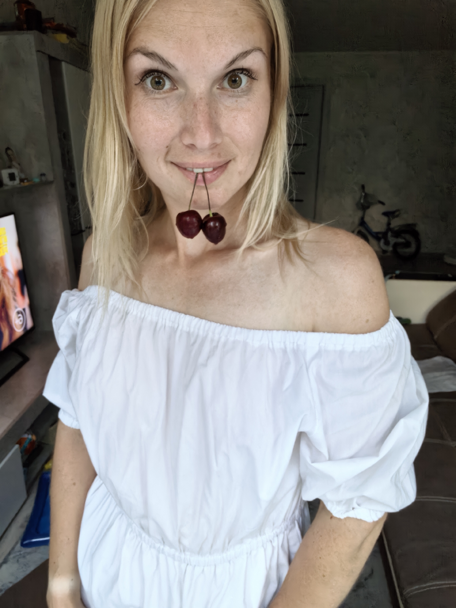 The image shows a young woman with blonde hair and freckles, wearing a white off-the-shoulder dress. She is holding two cherries between her teeth and looking at the camera. She is indoors, in what appears to be a living room or lounge area. In the background, there is a television set, some furniture, and a bicycle. The image does not provide any information that would indicate its location in Sterlitamak, Russia.