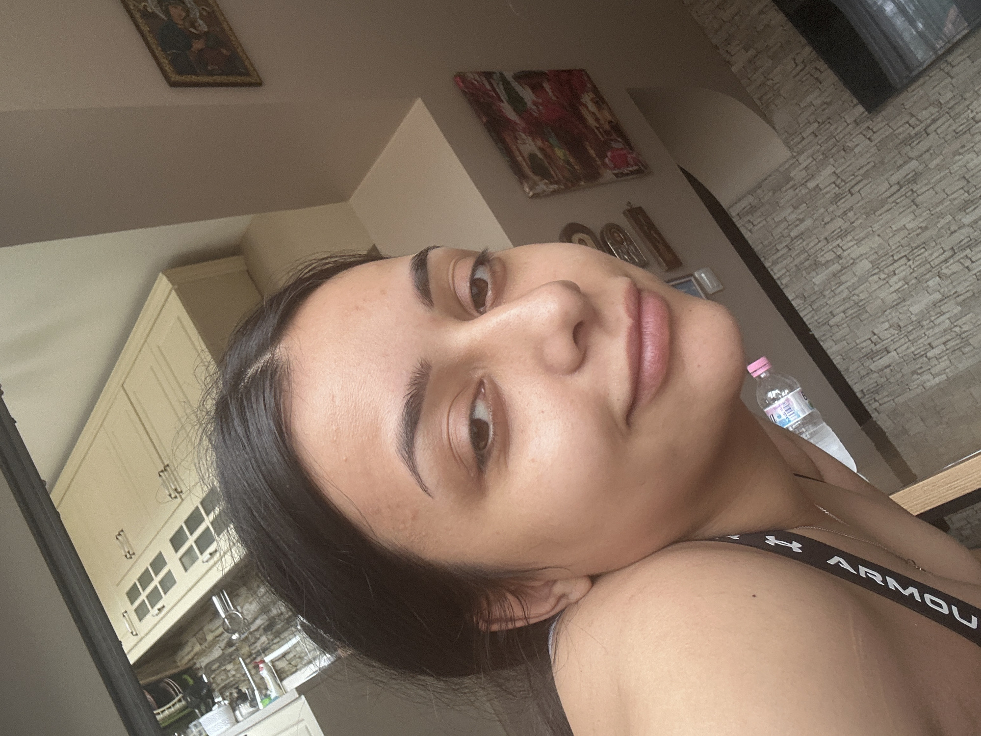 A young woman is captured in a close-up, tilted selfie shot indoors. She has dark hair, dark, well-defined eyebrows, and is looking directly towards the camera with a relaxed, slight smile. Her skin appears clear and natural. She is wearing a dark top, the strap of which prominently displays the white text "ARMOUR" along with an "H" logo, indicating the brand Under Armour. A thin gold chain necklace is also visible around her neck.
The background reveals an indoor residence, likely in Shumen, Bulgaria. To the left of the woman, a kitchen area is visible with light-colored, cream-toned upper cabinets featuring glass-paned doors and metallic handles. Below the cabinets, a sink area with some items like bottles and utensils can be discerned. To her right, a textured accent wall constructed from light-colored, stacked stone or brick adds architectural detail.
Above her head, on a plain light-colored wall, hang two framed artworks: one appears to be a traditional religious icon, and another is a more abstract painting with discernible red and dark brushstrokes. Other small decorative items are also present on the wall. A clear plastic water bottle with a pink cap is casually placed near her shoulder. The scene is brightly lit by natural light, suggesting it is daytime, and conveys a casual, relaxed atmosphere.