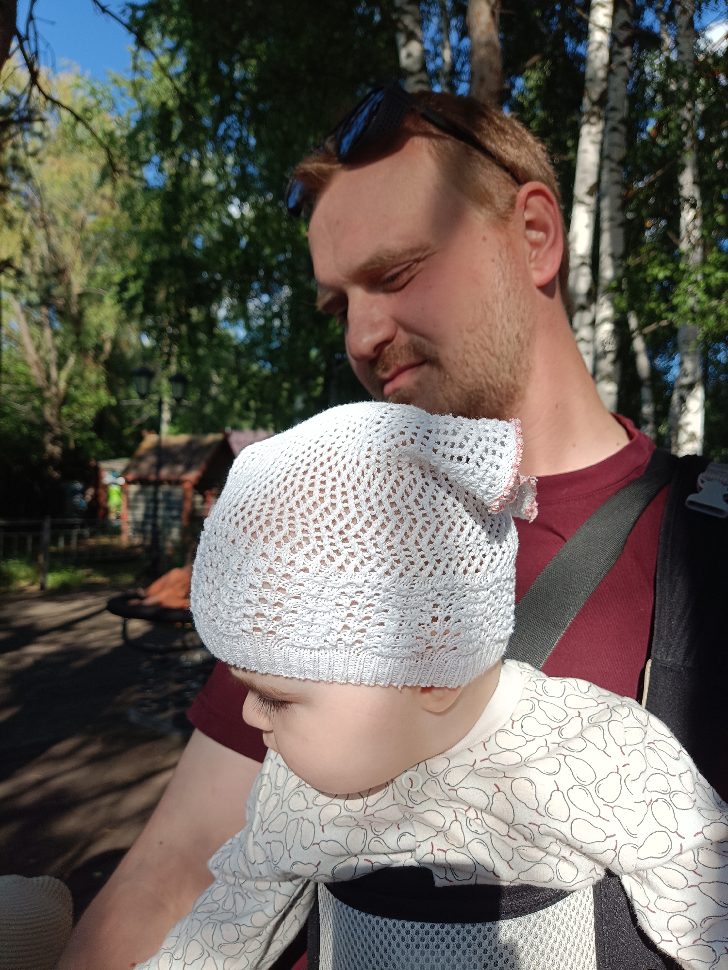 In the city of Chaikovsky, Russia, a man with reddish-blond hair and stubble is holding a baby in a carrier. The man is wearing a dark red t-shirt and sunglasses on his head. The baby is wearing a white knitted hat and a white long-sleeved shirt with a pear pattern. They are outdoors, with green trees and a clear blue sky in the background. The man is looking down at the baby. A small wooden building is visible in the background, along with a lamppost.