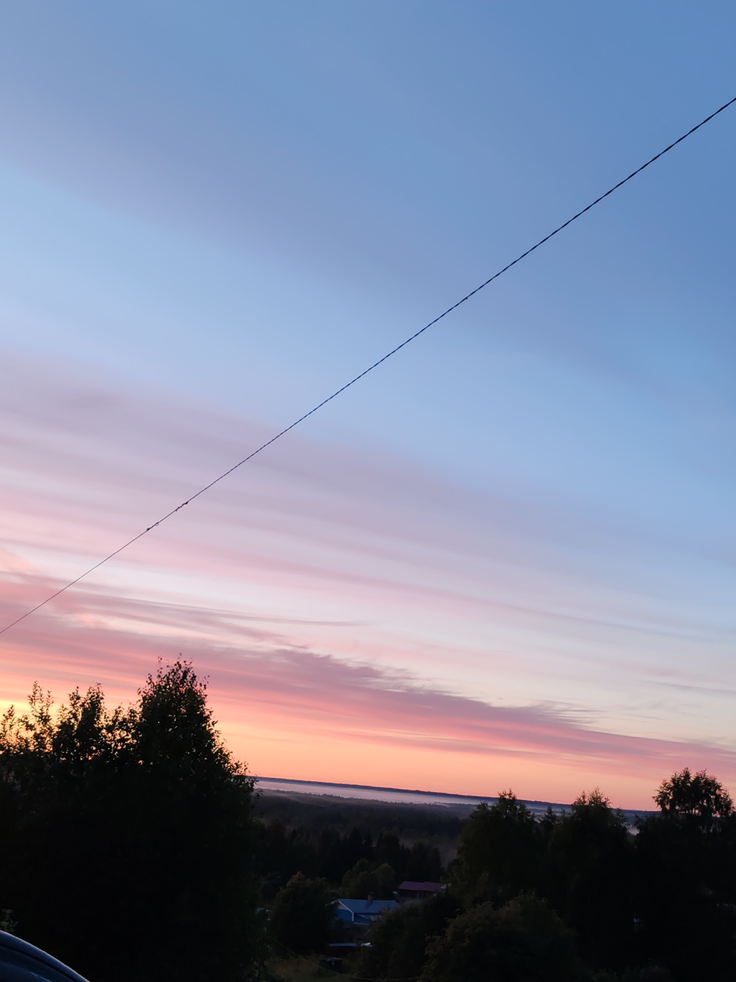 The image captures a serene sunset scene over a rural landscape in or near Chukhlem, Russia. The sky is a canvas of soft gradients, transitioning from a clear, pale blue at the top to delicate hues of pink and orange near the horizon. Wispy clouds streak horizontally across the sky, catching the light of the setting sun. A single, thin power line bisects the sky diagonally from the top left to the center right. Silhouetted against the colorful sky are dark green trees and foliage, forming a dense line across the lower third of the image. In the distance, the landscape appears to be a forest, with a light mist or fog settling low on the horizon. A few small buildings, including a blue-roofed structure, are visible nestled amongst the trees in the middle ground. The foreground shows the edge of a dark-colored vehicle. No people are visible in the frame.