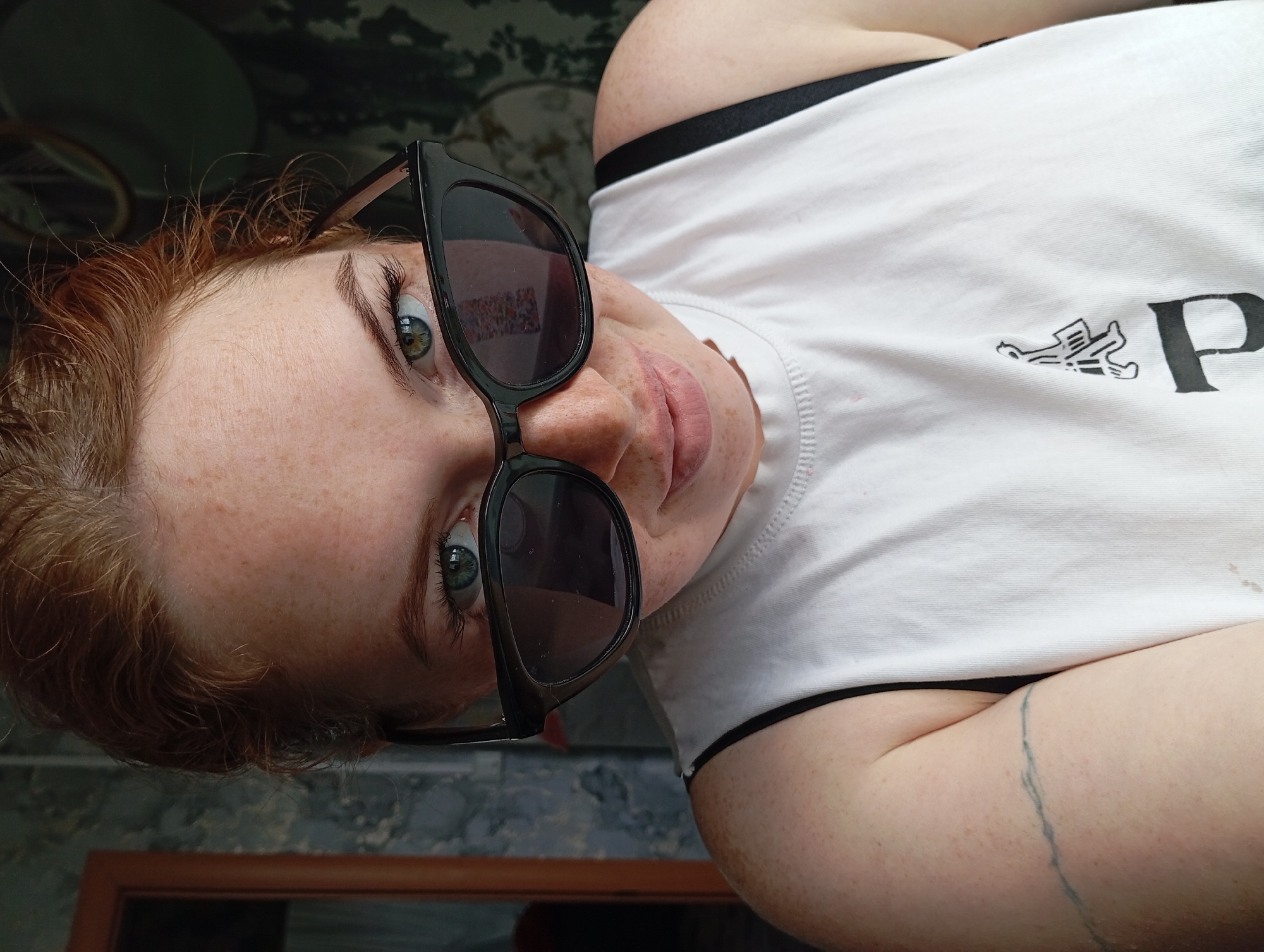 A fair-skinned person with red hair and freckles is wearing black sunglasses and a white t-shirt with black lettering. They are looking directly at the camera from a low angle. The background appears to be an indoor scene with patterned wallpaper and a portion of a wooden frame. The location is unknown.
