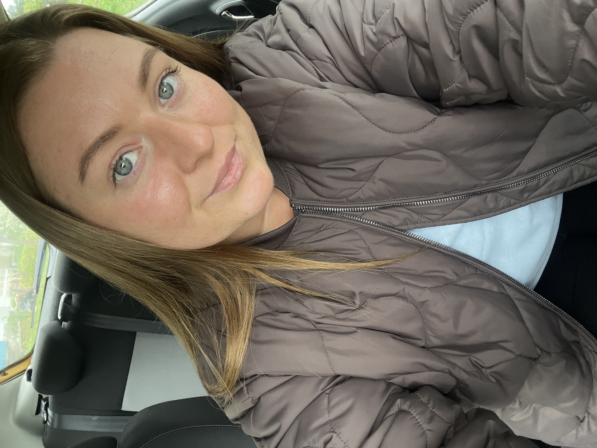 A woman with light brown hair and light eyes is taking a selfie in the front seat of a car. She is wearing a quilted brown jacket over a white shirt. The car's interior is visible, including the black seats and seatbelt. Outside the car window, a blurred cityscape can be seen. The location is Раздолье, Russia.