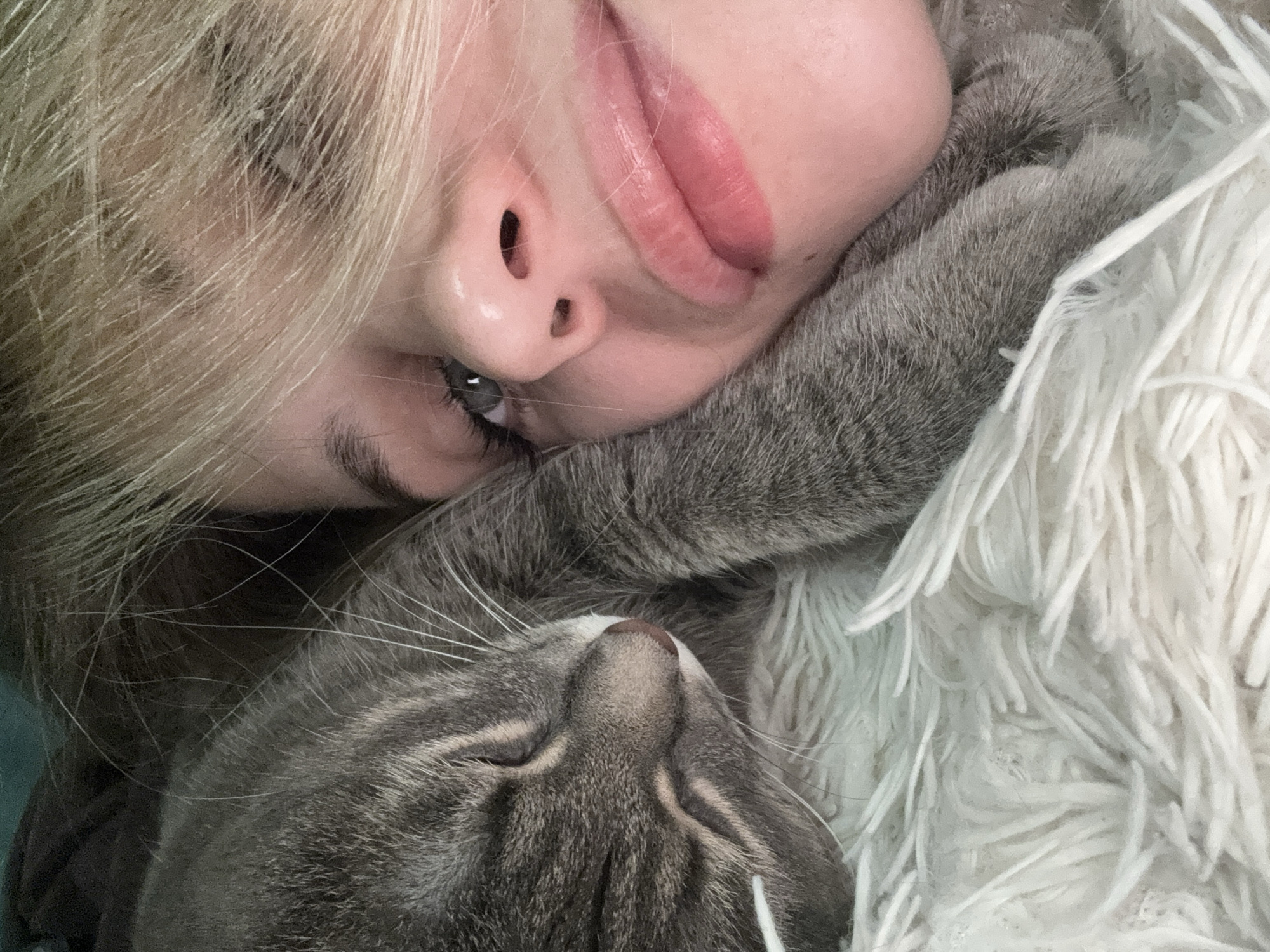 A blonde woman and a grey tabby cat are lying down together, likely indoors, on a white, shag-like blanket or rug. The woman's face is visible, and she appears to be resting her head on the cat. The cat is also lying down, with its head near the woman's face. The location cannot be determined from the image.