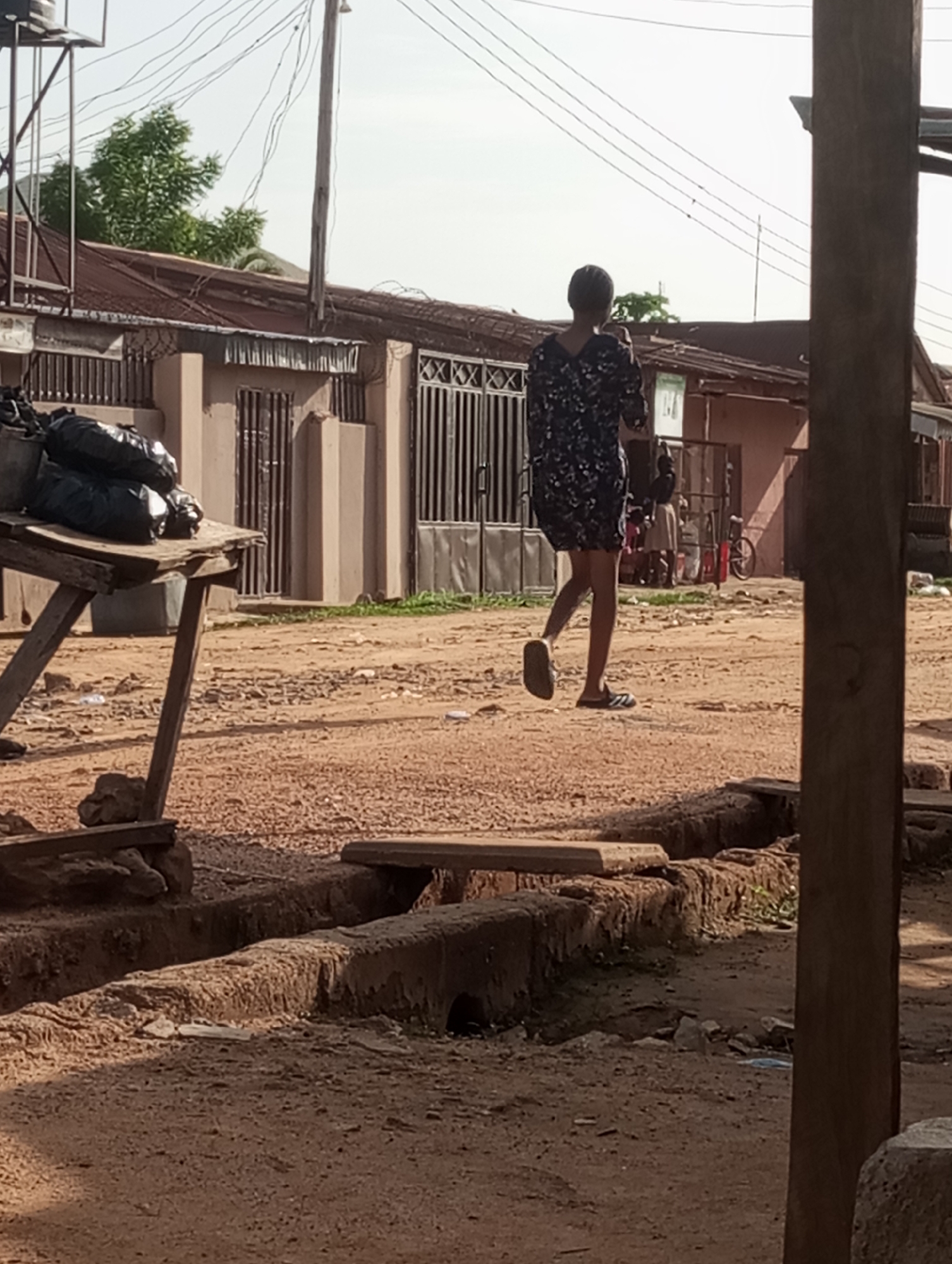 A person in a patterned dress and sandals is walking down a dirt road in Narayi, Nigeria. Buildings with barred windows are on either side of the road. A makeshift table loaded with black bags stands to the left. An open drainage ditch runs along the road's edge. Other people and a bicycle are visible in the background near a shop. Overhead are numerous power lines. A large wooden pole is visible to the right.