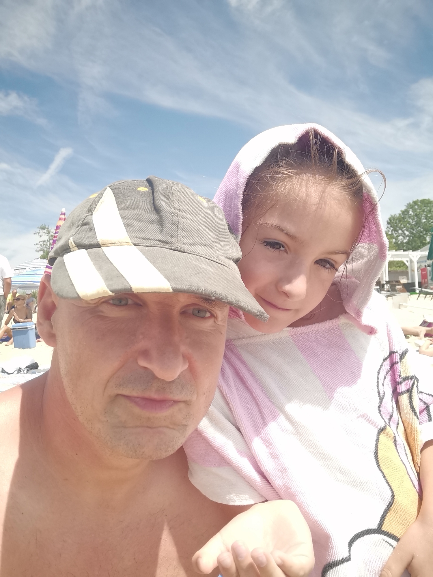 The image is a selfie taken outdoors on a sunny day, likely at a beach in Kranevo, Bulgaria. The foreground features two subjects: a man wearing a grey baseball cap and a young girl wrapped in a pink and white striped towel with a hood. The man is in the lower left corner, looking directly at the camera with a neutral expression. The girl is positioned behind and to the right of the man, with her face visible as she looks slightly off-camera with a gentle smile. Her hair appears damp, suggesting they may have been swimming.
In the background, the scene depicts a beach environment. There are beach umbrellas, lounge chairs, and other beachgoers, indicating a lively atmosphere. The sky is blue with scattered white clouds. The lighting suggests it is daytime, likely late morning or early afternoon. The overall mood appears relaxed and familial, as if capturing a moment of leisure during a vacation. No visible text is present in the image.