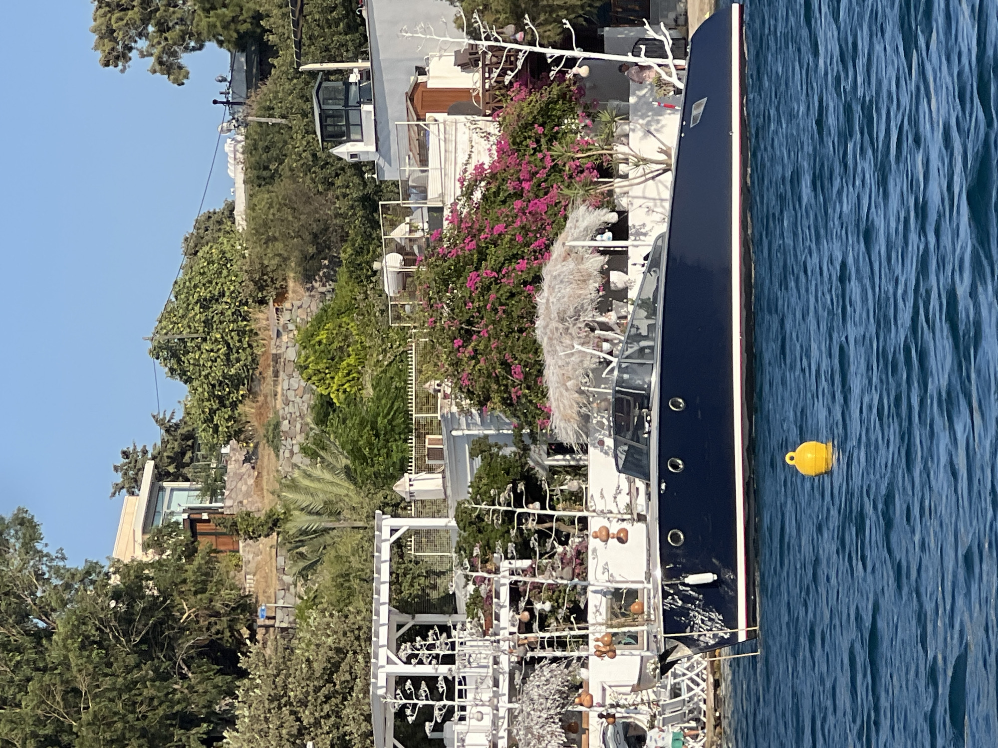 A dark blue yacht with a white stripe along its waterline is docked in blue water. In the background, a white building with white pergolas and lush green trees with pink flowers are visible. A yellow buoy floats in the water to the left of the yacht. The scene is illuminated by bright sunlight, casting shadows. The sky is clear and blue.