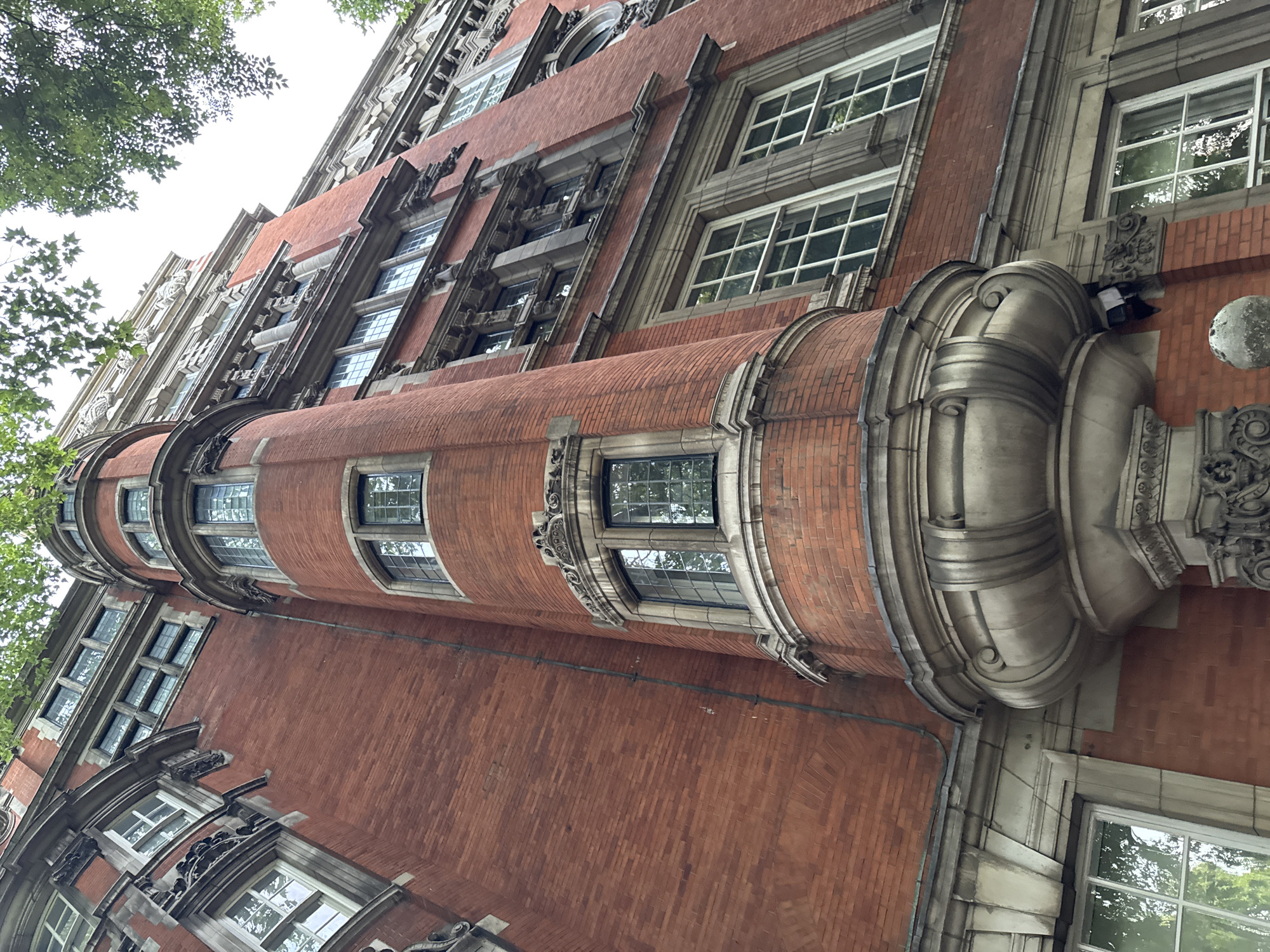 A low-angle, full-shot photograph captures the facade of a red brick building in London, United Kingdom, under a partially obscured sky by green tree leaves. The building exhibits classical architecture, with a prominent cylindrical tower on the left featuring ornate stone detailing around its windows and base. The tower's curved brickwork extends upwards, broken by a series of arched windows. To the right of the tower, the building's facade continues with multiple stories of windows, characterized by white frames and mullions, interspersed with decorative stone cornices and architectural embellishments. The overall impression is one of grandeur and historical significance, likely a public institution or older commercial building. No people or specific activities are visible in the frame.