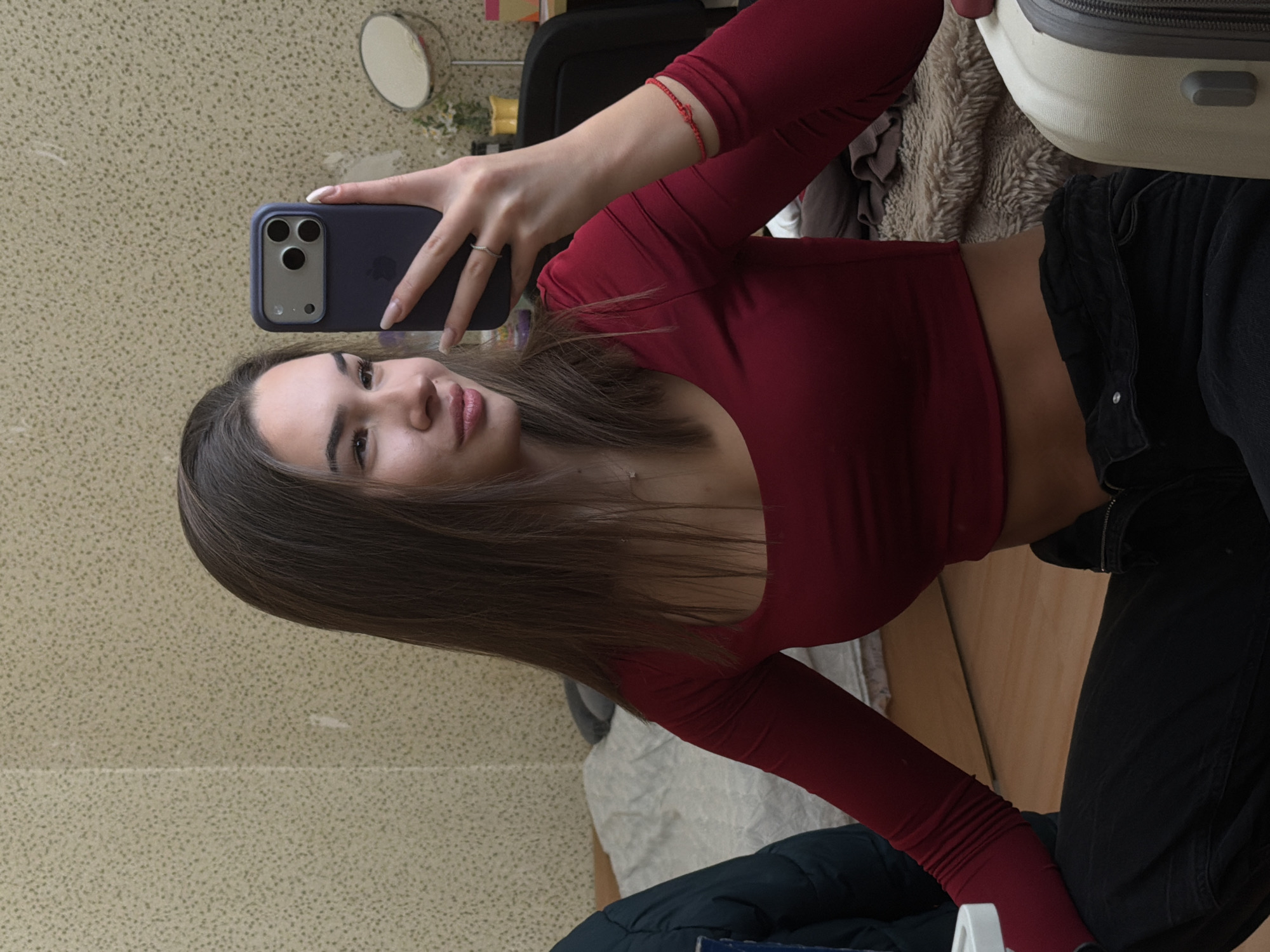A young female is depicted in an indoor setting, presumed to be a residential room in Sofia, Bulgaria. She is seated or kneeling on a light brown wooden floor, facing towards her right and looking into a reflective surface while holding a dark blue iPhone with a triple-lens camera in her left hand to capture a selfie.
The female has long, straight, brown hair, and wears a dark red, long-sleeved crop top that exposes her midriff, paired with black trousers. A red string bracelet is visible on her right wrist, a silver ring on her left ring finger, and a thin silver necklace around her neck. Her fingernails are manicured.
The background consists of a light-colored, textured wallpaper with a speckled pattern, exhibiting minor wear and discoloration. To the left, a section of a bed with a light-colored sheet and a dark grey pillow is visible, alongside a dark teal puffer jacket draped over an unseen object. To the right, a light-colored hardshell suitcase with a grey zipper is present in the lower foreground. Further in the background, a dark office chair, a small round magnifying mirror on a stand, a vase containing white flowers, and a yellow decorative object are discernible on a surface, likely a desk or dresser. A furry, light brown blanket or throw is also partially visible on the floor or furniture in the lower right background.