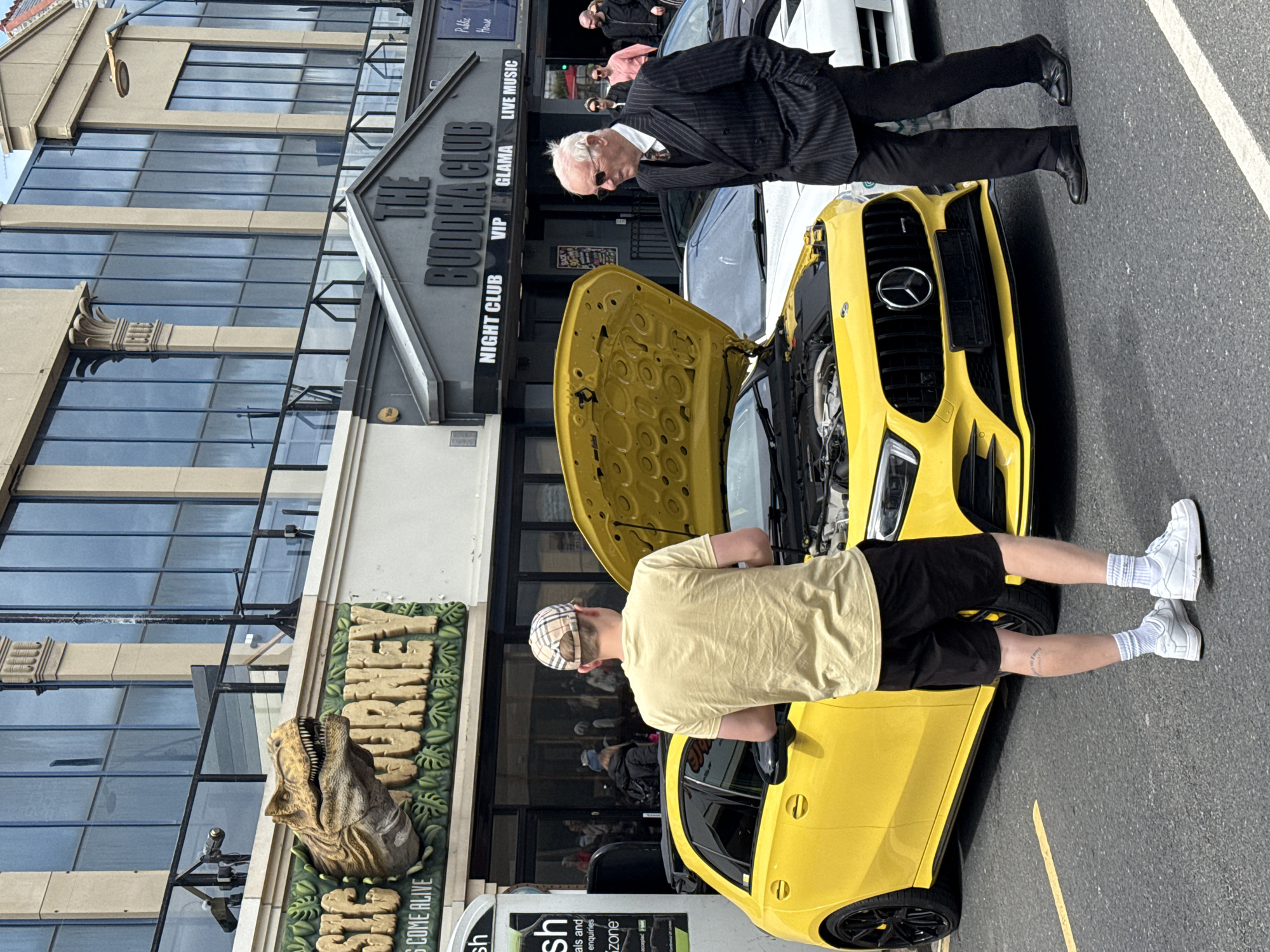 The image, taken in Great Yarmouth, UK, shows two people examining a yellow Mercedes car with its hood open. One person, an older man in a pinstripe suit and sunglasses, is leaning over the front of the car. The second person, a younger individual with a checkered cap, tan t-shirt, and black shorts, stands next to the car's open door. Behind the car is a building with a sign that says "The Buddha Club" with labels for "Night Club," "VIP," "Glama," and "Live Music." To the left, there's a sign for "Jurassic Journey" with a dinosaur head protruding from the sign. Behind "Jurassic Journey" is a building with large windows. The scene suggests the car may have had a mechanical issue. The photo is taken on a street with asphalt.
