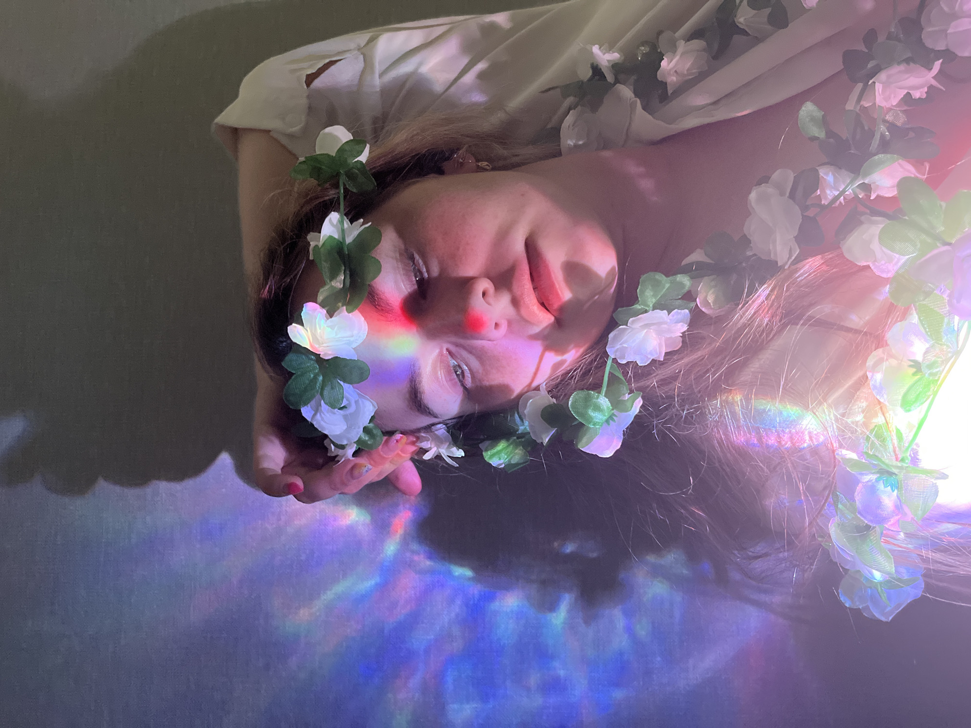 A woman with fair skin and blonde hair lies on a surface, surrounded by artificial white flowers with green leaves. She is wearing a light-colored top. Her eyes are partially closed, and a faint red tint appears on her cheeks and the bridge of her nose. A prism-like effect of diffused colors – predominantly blues, purples, and hints of red – are cast across the surface and the surrounding areas, creating an ethereal ambiance. Her hand is partially visible, resting on the surface near her face. The background is a soft, neutral tone. This image does not allow for the determination of a specific location like the city of Chaikovsky, Russia.