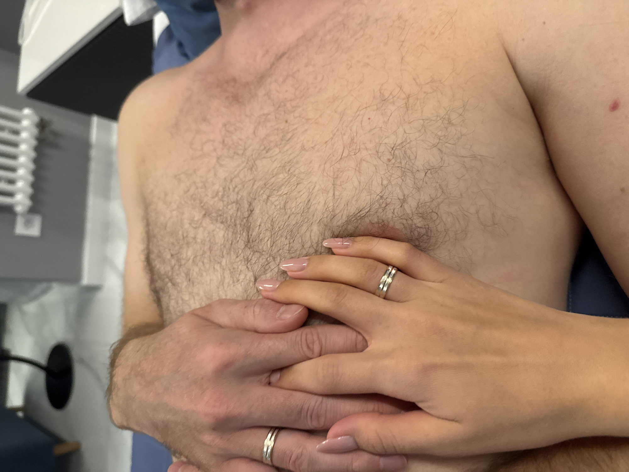 A close-up shot depicts the upper torso of a fair-skinned, hairy man, with his chest and shoulders visible. Overlapping his chest are the hands of another fair-skinned person, presumably female, adorned with a silver ring on the ring finger of the right hand. The man also wears a matching silver ring on his left ring finger. The nails are manicured in a light pink shade. The backdrop includes a partial view of what seems to be a radiator and a blue surface, possibly a sheet or blanket. The location is ambiguous.
