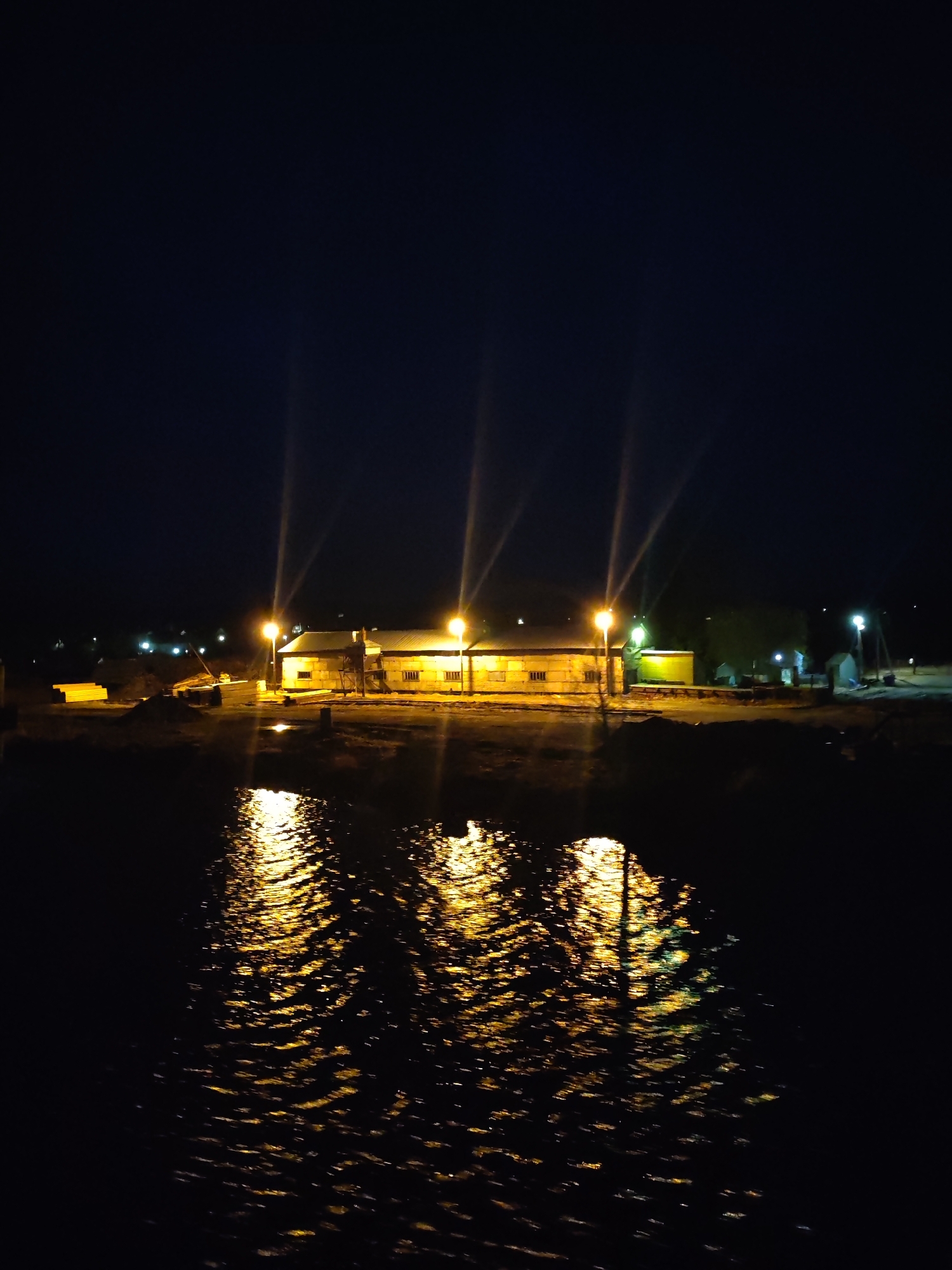 A nighttime scene shows a body of water in the foreground reflecting the lights from buildings and street lamps on the opposite shore. The main building appears to be a long, low structure illuminated by at least four light poles. The reflections in the water create a distorted pattern of gold and light. Additional buildings and streetlights are visible in the background, suggesting a settled area. The sky is dark. The overall impression is a combination of darkness and localized illumination.