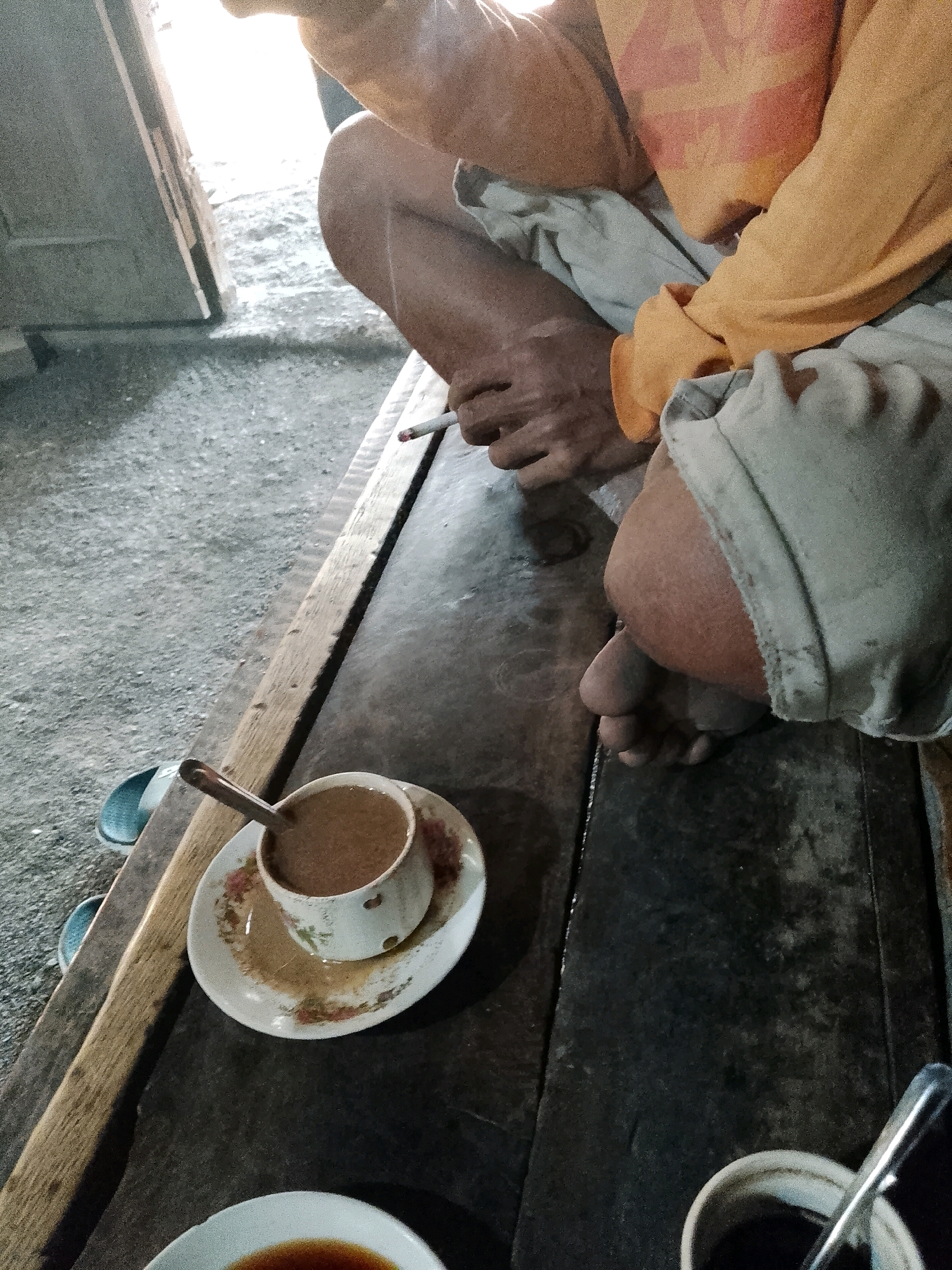 A person, seated on a wooden bench, is smoking a cigarette and holding a cup of coffee. The coffee cup is white with a floral pattern and rests on a matching saucer. A spoon is in the cup. The person is wearing an orange long-sleeved shirt and light-colored pants. Their legs are crossed, and their bare feet are visible. The background shows a concrete floor and a doorway. The scene appears to be outdoors, possibly in a rural setting in Wadang, Indonesia, given the informal arrangement and simple furnishings.