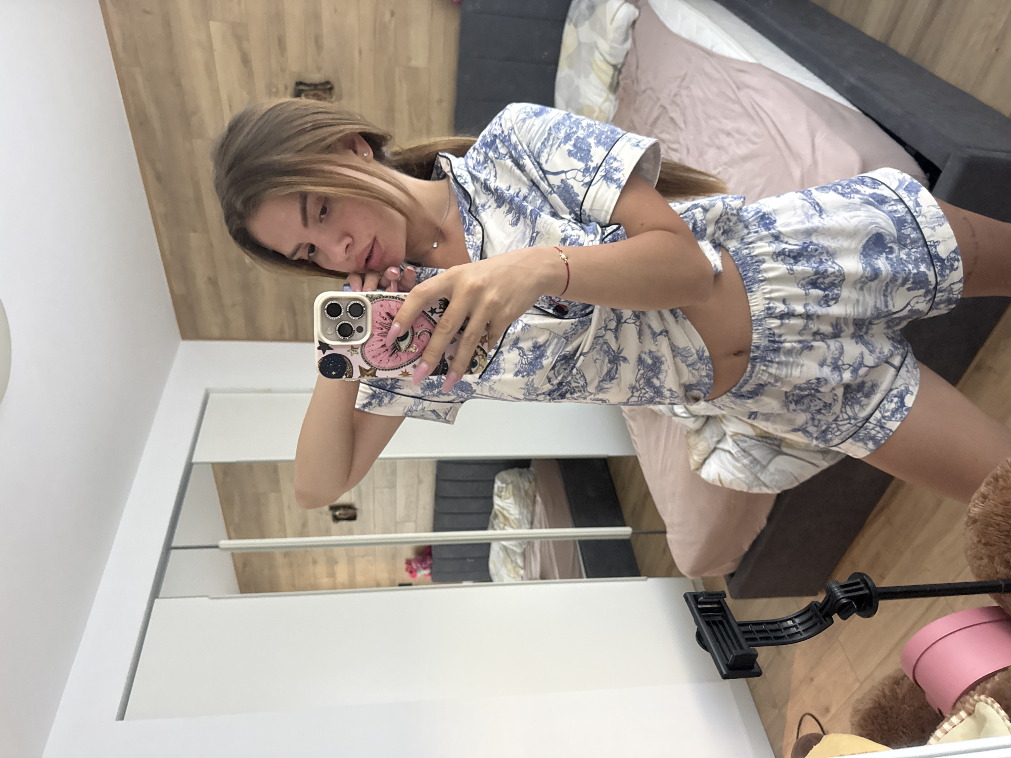 A young woman is depicted taking a mirror selfie in a bedroom setting. The image is rotated 90 degrees clockwise. She has long, light brown hair styled in a half-up ponytail and is looking directly at her smartphone, which she holds up to capture her reflection. Her expression is calm and relaxed, with her lips slightly parted. She is wearing a short-sleeved pajama set, white with blue toile-style patterns featuring trees and natural scenes. The top is slightly unbuttoned, revealing a sliver of her midriff, and she wears a silver necklace with a small pendant. A thin red string bracelet with a small gold charm adorns her right wrist, and her nails are painted a light pink or nude shade.
The smartphone she holds has a distinctive light-colored case adorned with various designs, including stars and a prominent eye-like symbol.
The reflection in the mirror provides a glimpse of the bedroom. A bed with a dark grey headboard and a light pink duvet or comforter is visible. The room has light wooden floorboards. To the left of the bed, a white wall is reflected. On the floor in the reflected image, a pink cylindrical hatbox or container is seen, next to what appears to be a large, fluffy, light brown object, possibly a teddy bear or blanket. A black, articulated phone stand or tripod arm is also present on the floor. The left edge of the mirror's reflection shows part of a white wardrobe or cabinet with reflective surfaces.
The overall scene suggests a private, domestic moment, likely during the daytime due to the bright ambient light. No legible text is visible in the image.
