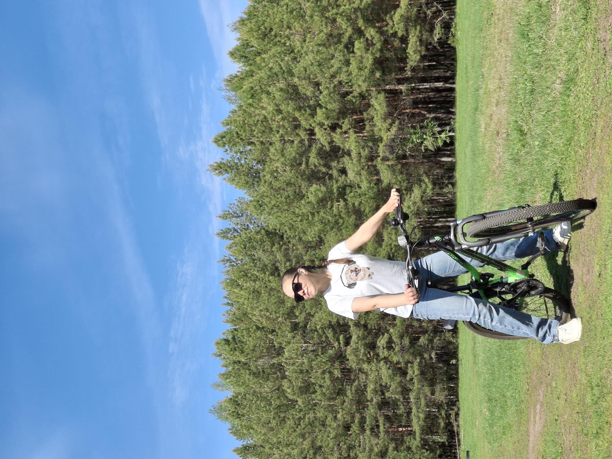 A young woman with sunglasses stands next to a green and black mountain bike in a grassy area bordered by a dense green forest on one side and a bright blue sky on the other. The location is Chaikovsky, Russia. The woman wears a white t-shirt, blue jeans, and white sneakers. She is holding the handlebars of the bike with her right hand.