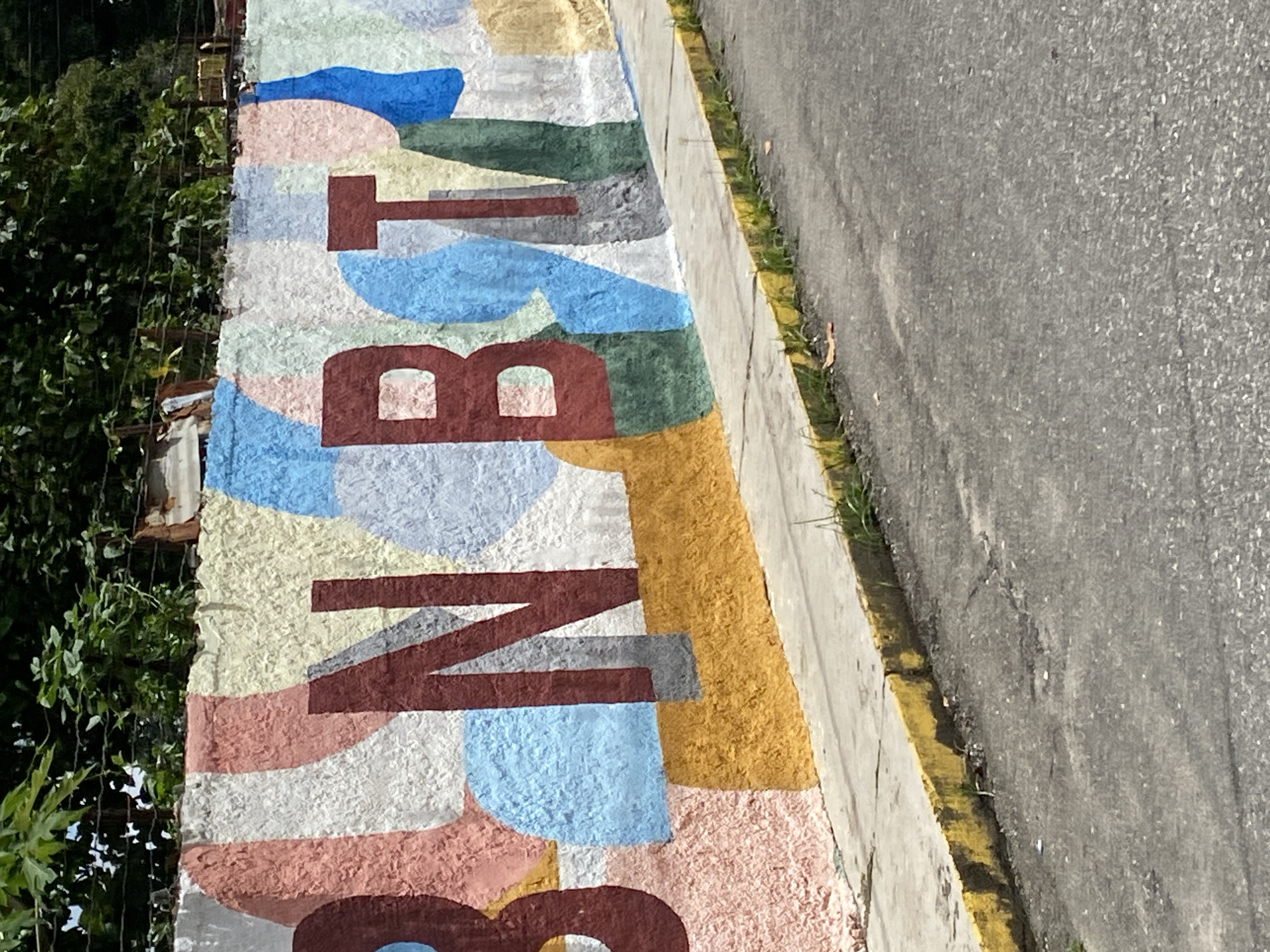 The image captures a portion of a colorful mural along a sidewalk in Tinaquillo, Venezuela. The mural displays large, block letters reading "BARRIO". The backdrop consists of various irregularly shaped patches of color, including blues, greens, yellows, reds, and whites. The mural is located on a wall bordering a street, with some greenery visible on the left side of the frame. The sidewalk borders a street with a yellow curb.