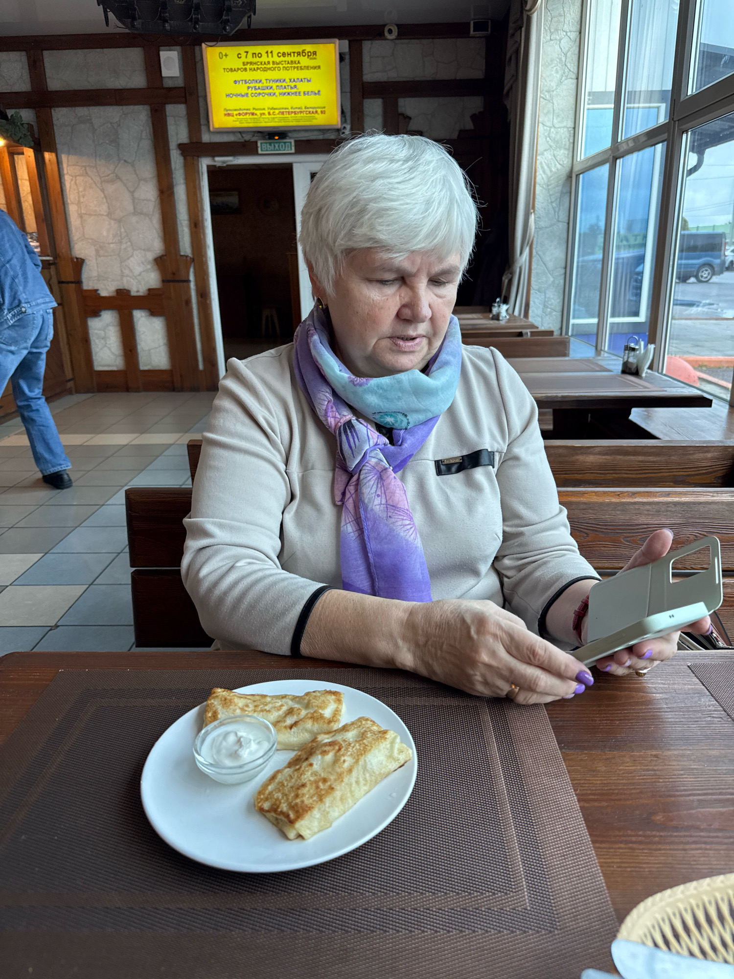 A middle-aged woman with short, white hair is sitting at a wooden table in a restaurant and looking at a smartphone. She is wearing a light-colored sweater and a purple and blue patterned scarf. In front of her on the table is a white plate with two rolled-up crepes and a small bowl of white sauce. The restaurant has wooden booths and large windows. In the background, a sign is visible with Russian text that appears to be advertising an exhibition and stating dates in September. The flooring is a tile pattern of light and dark squares. Another person's legs in blue jeans and dark shoes are visible walking in the background to the left. The woman's fingernails are painted purple.