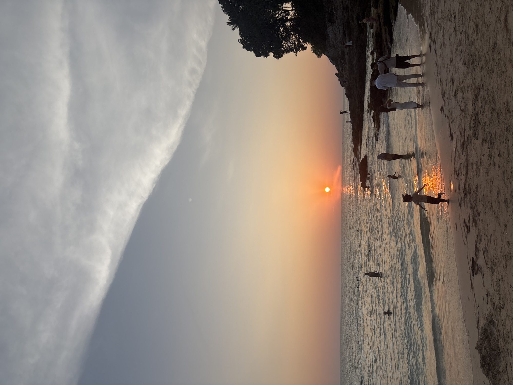 The image depicts a beach scene at sunset in Choeng Thale, Thailand. The sun is setting over the ocean, with its light reflecting on the water. Several people are on the beach and in the water, some standing near the shoreline and others wading further out. A rocky coastline with some trees is visible on the right side of the image. The sky is a gradient of colors, with gray clouds contrasting with the yellow and orange hues of the sunset.